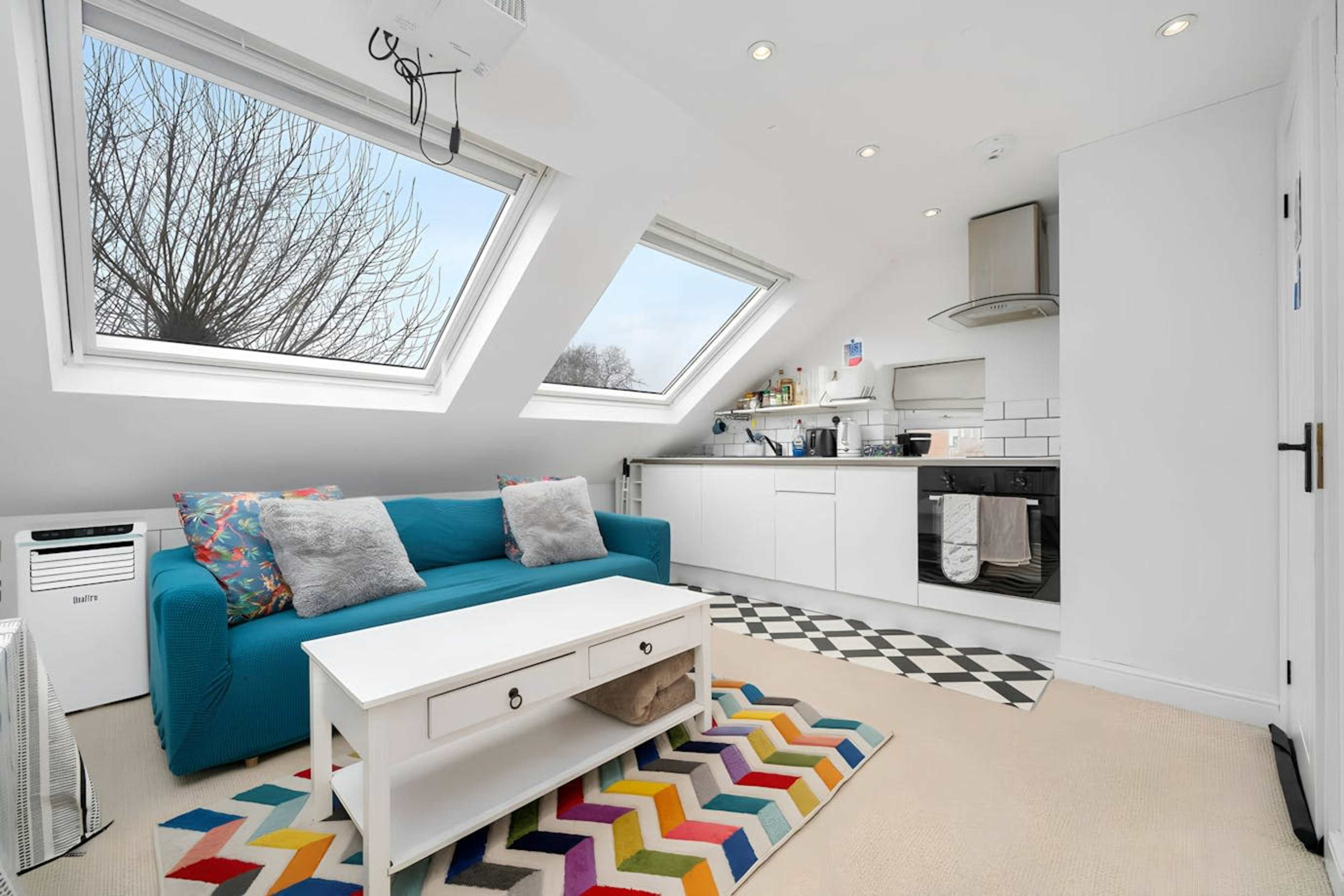 The image shows a small, modern living space with a blue couch, a white coffee table, and a kitchenette featuring skylight windows.