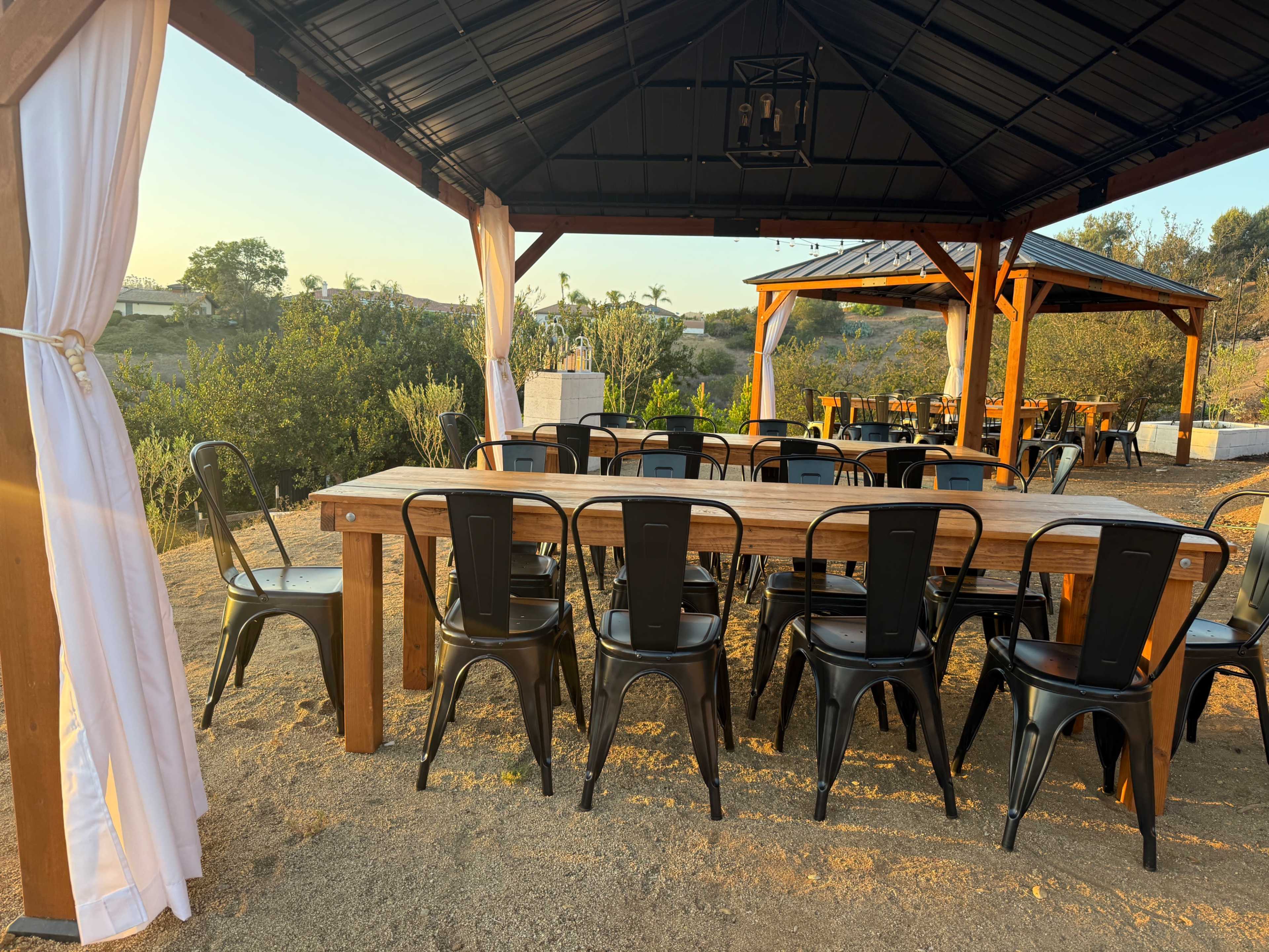 A spacious outdoor dining area features a long wooden table surrounded by black metal chairs under a gazebo structure at sunset.