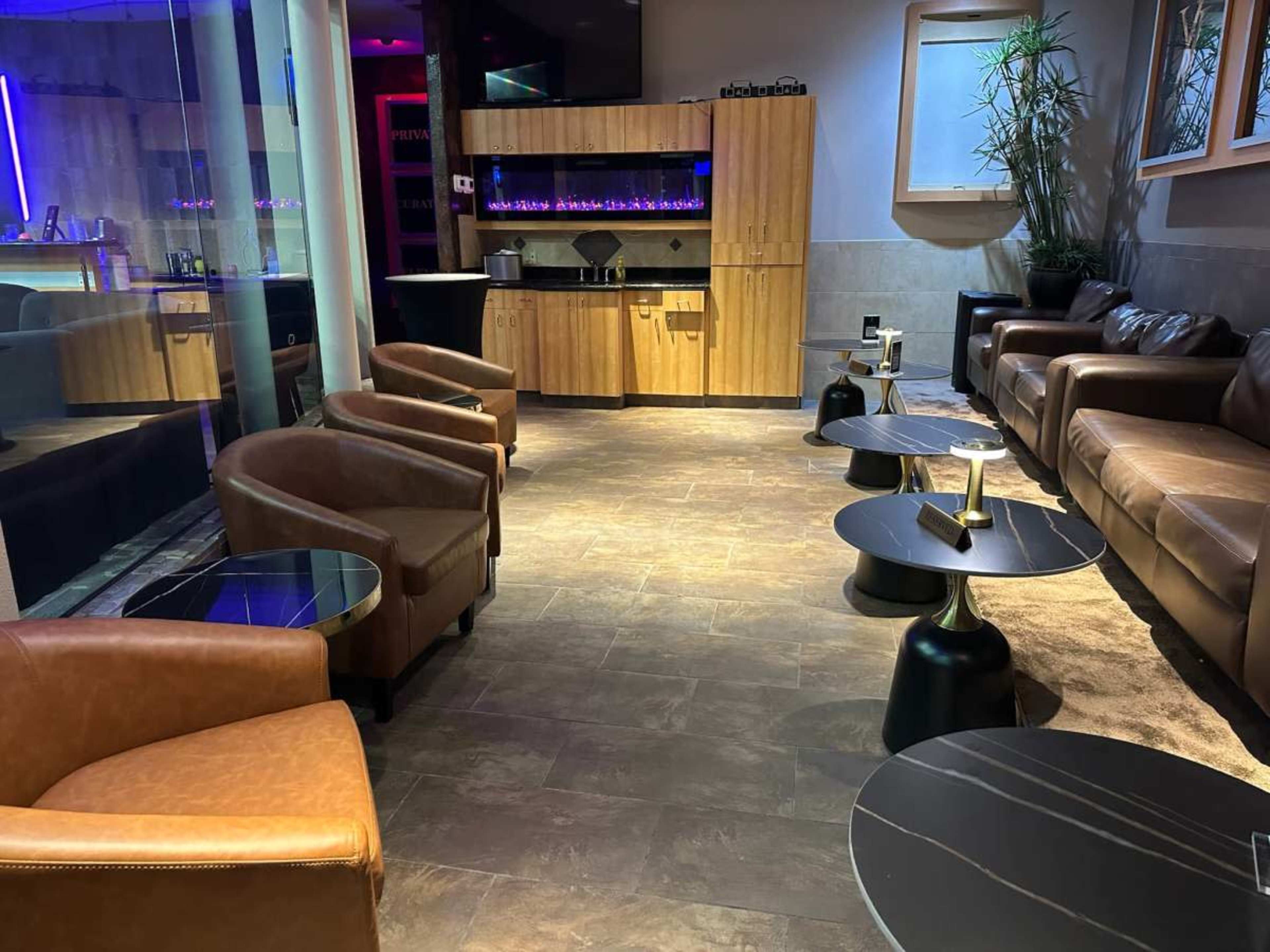The image shows a modern lounge area with leather armchairs and low tables, featuring soft lighting and a wooden service counter in the background.