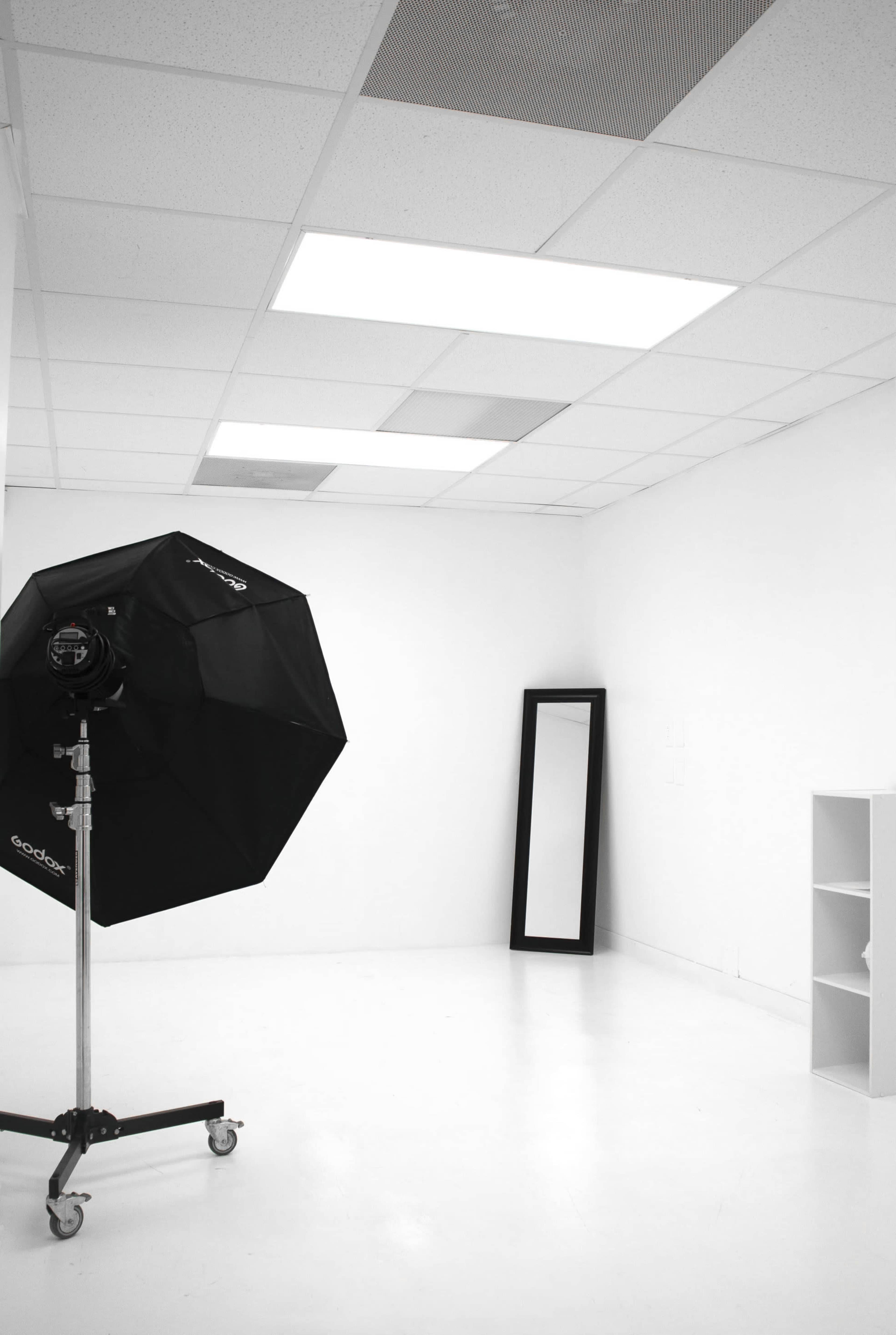 A minimalist white studio room with a large softbox light on a stand, a tall mirror, and a small shelving unit.