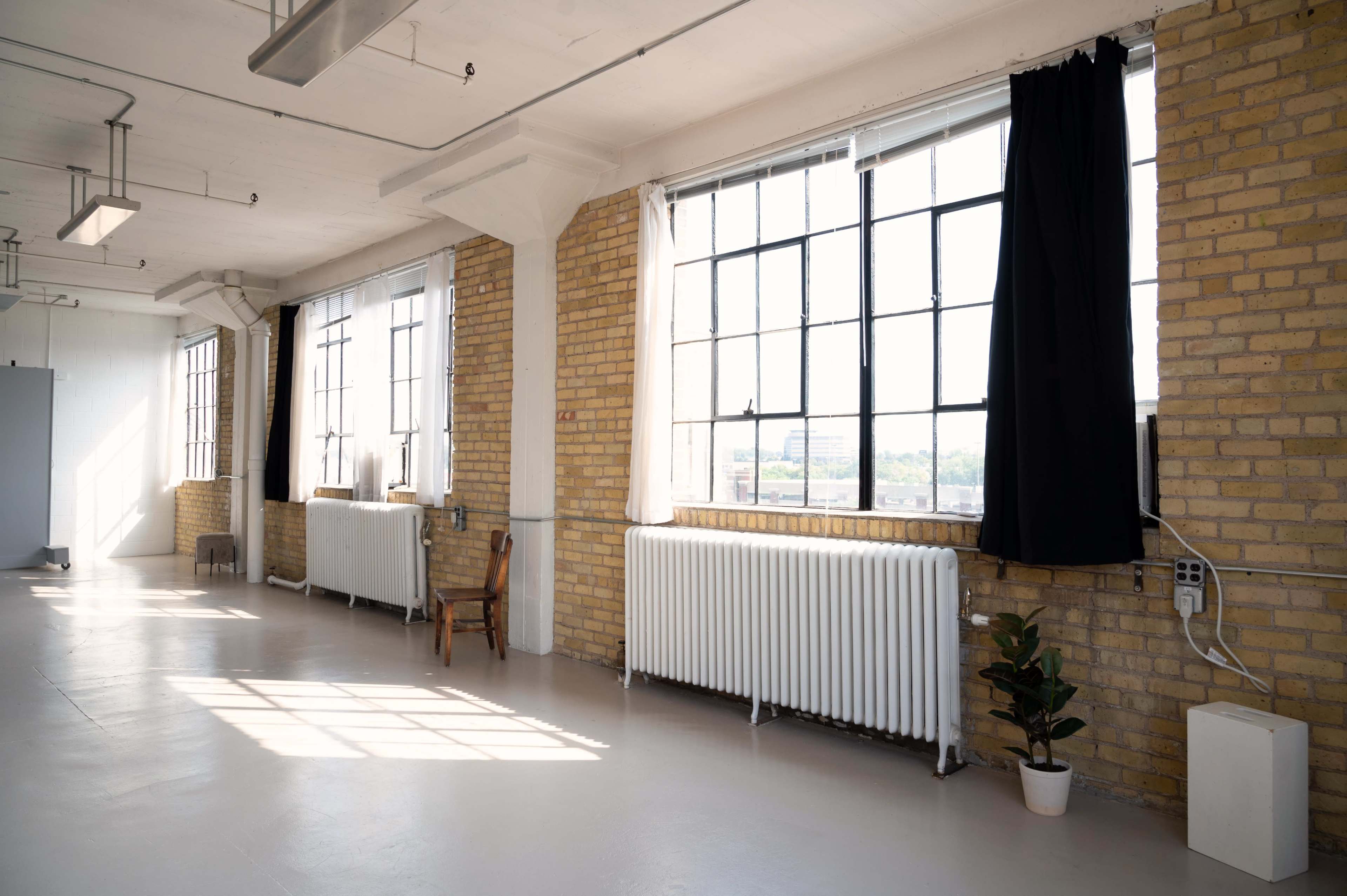 The image shows a bright, spacious room with large windows, exposed brick walls, and a few pieces of simple furniture.