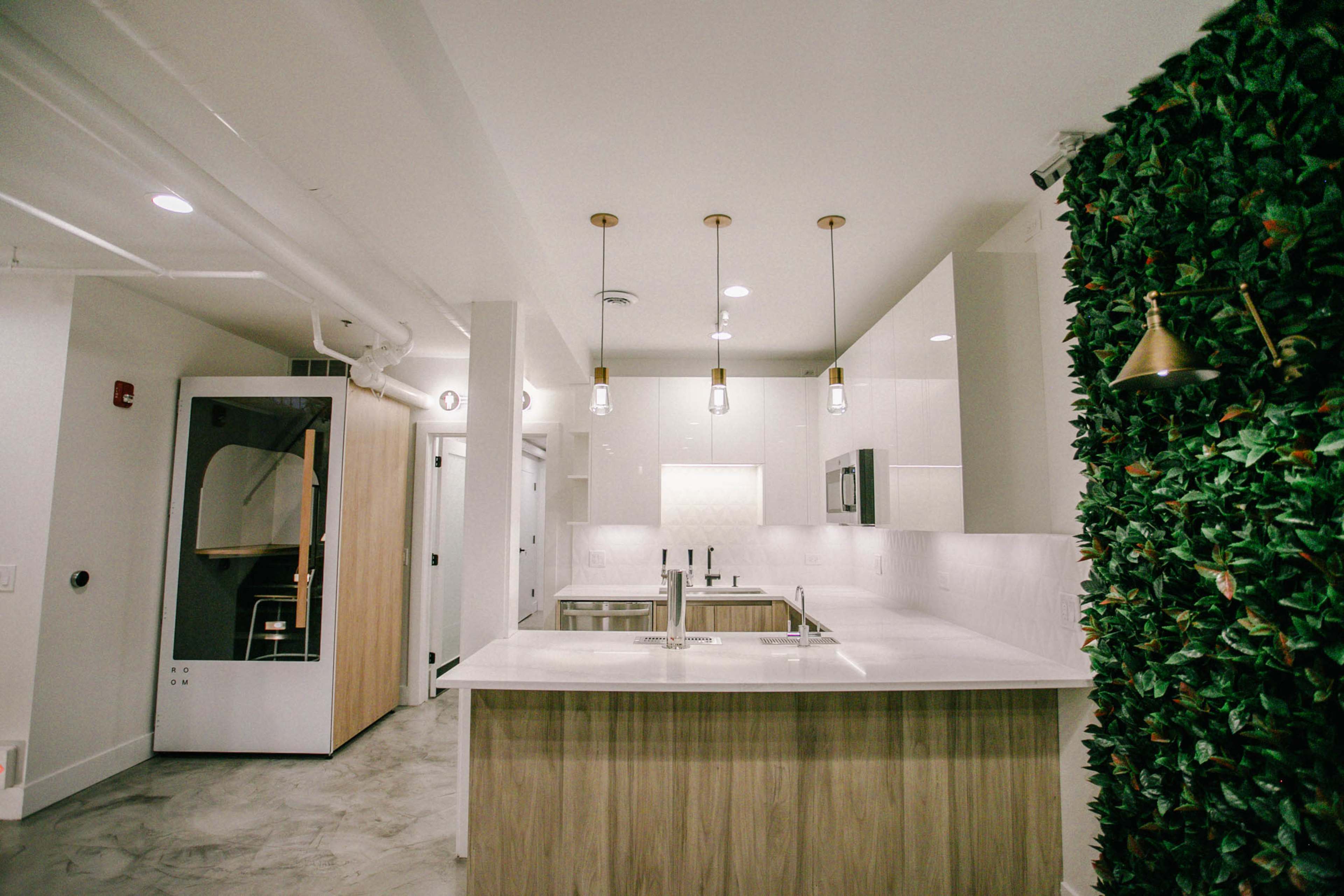 A modern kitchen featuring a central island with a white countertop, pendant lights, and a wall adorned with greenery.