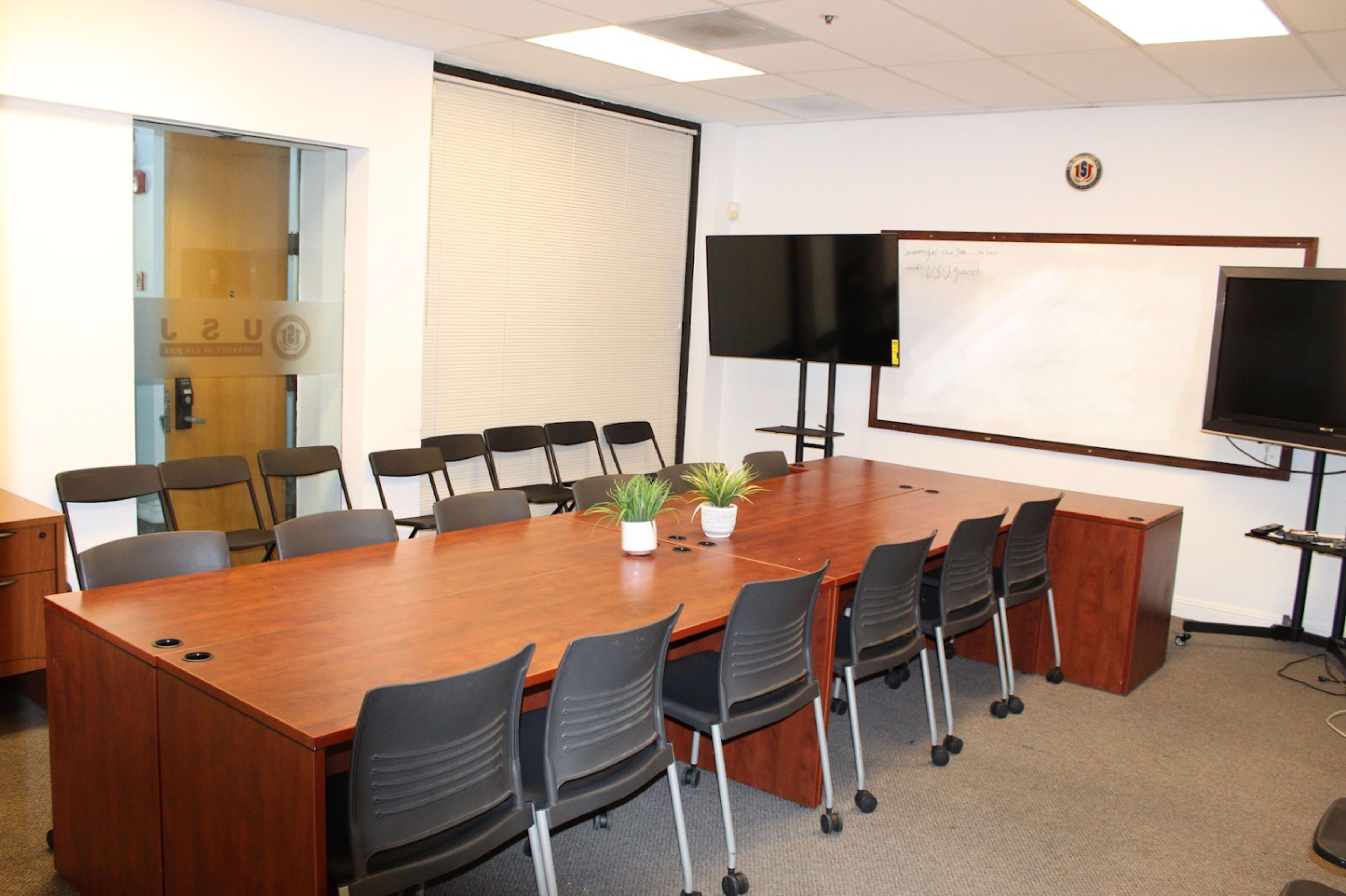 A conference room features a long wooden table surrounded by black chairs, with two large screens and a whiteboard on the walls.
