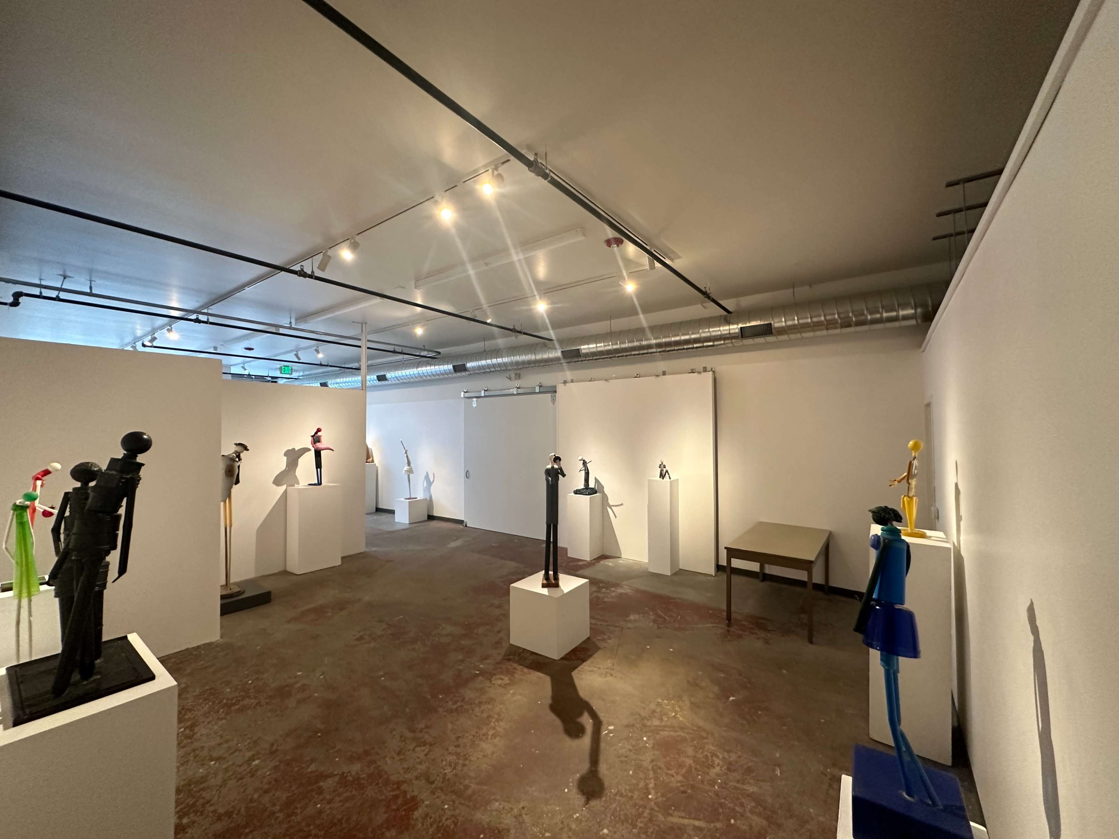 A minimalist art gallery features various sculptures displayed on white pedestals against a neutral wall.