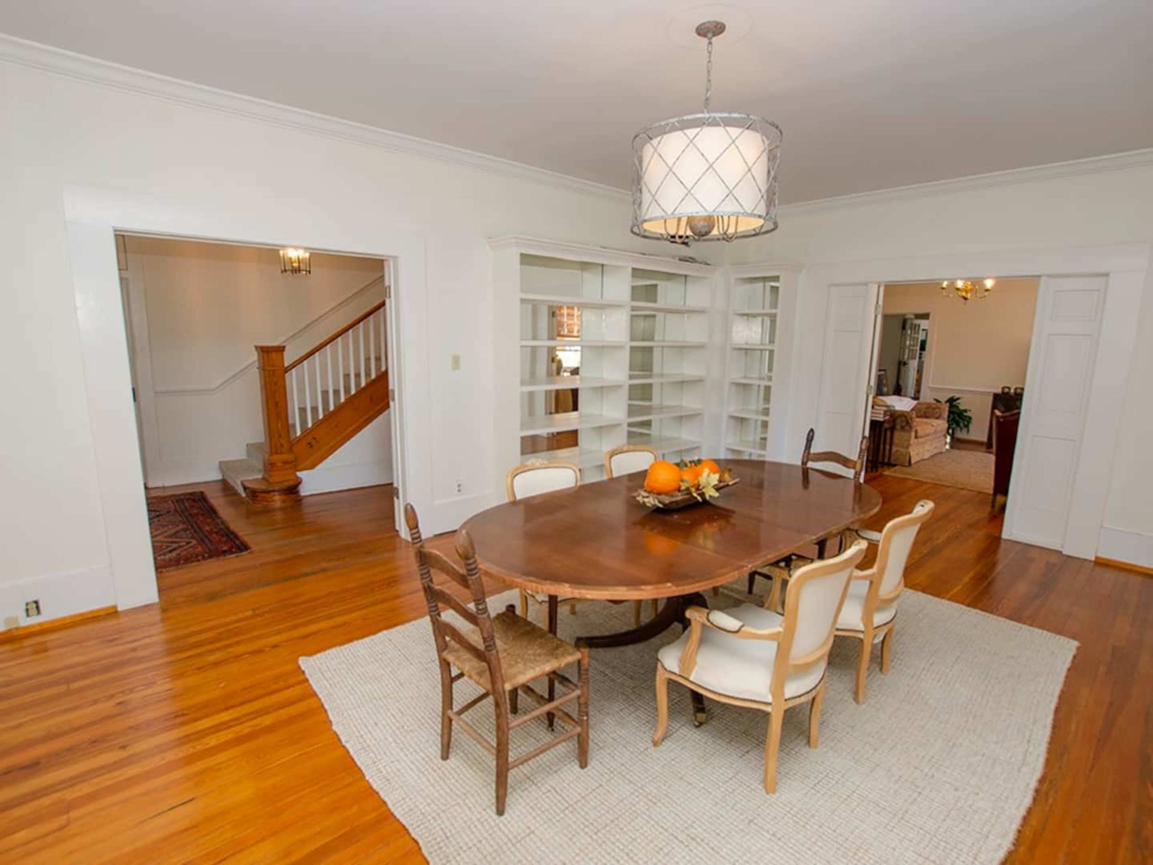 A spacious dining room features a large wooden table surrounded by eight chairs, with a decorative light fixture above and access to a staircase and adjoining rooms.
