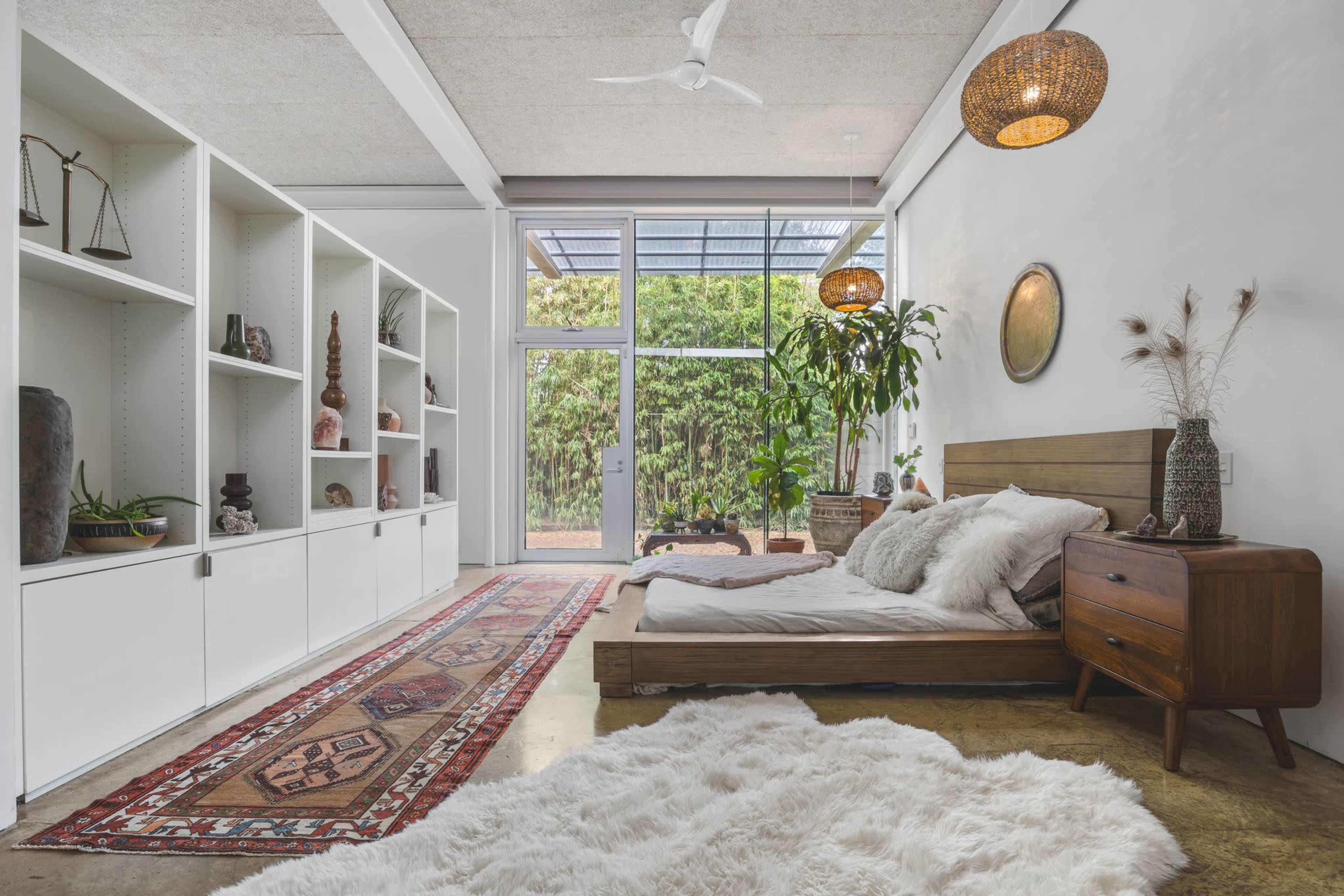 The image shows a modern bedroom featuring a low wooden bed, a decorative rug, and shelves displaying various objects, with large windows allowing natural light and views of greenery outside.