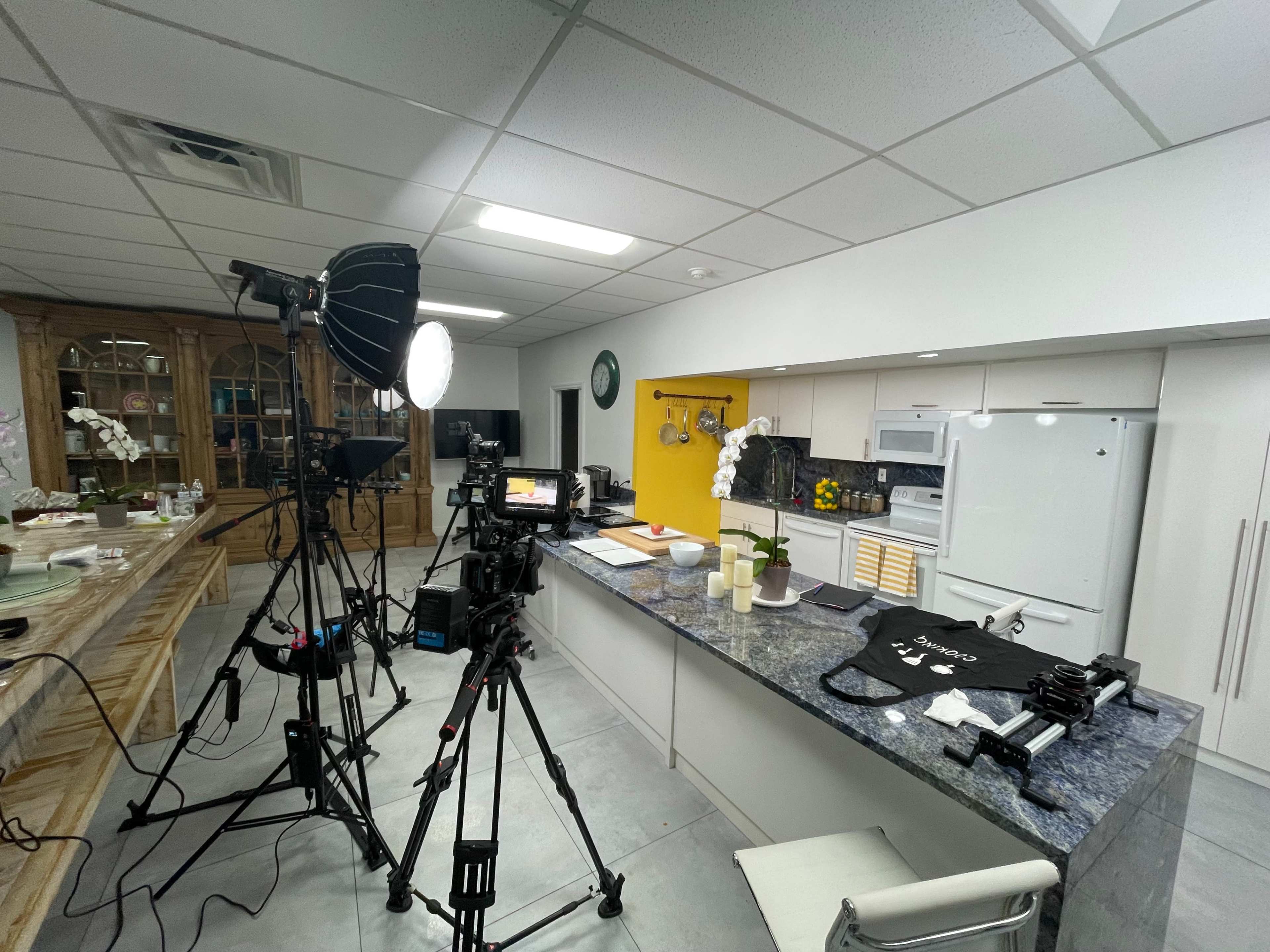 The image shows a well-equipped kitchen set up for filming, featuring cameras, lighting equipment, and various kitchen items on the counter.