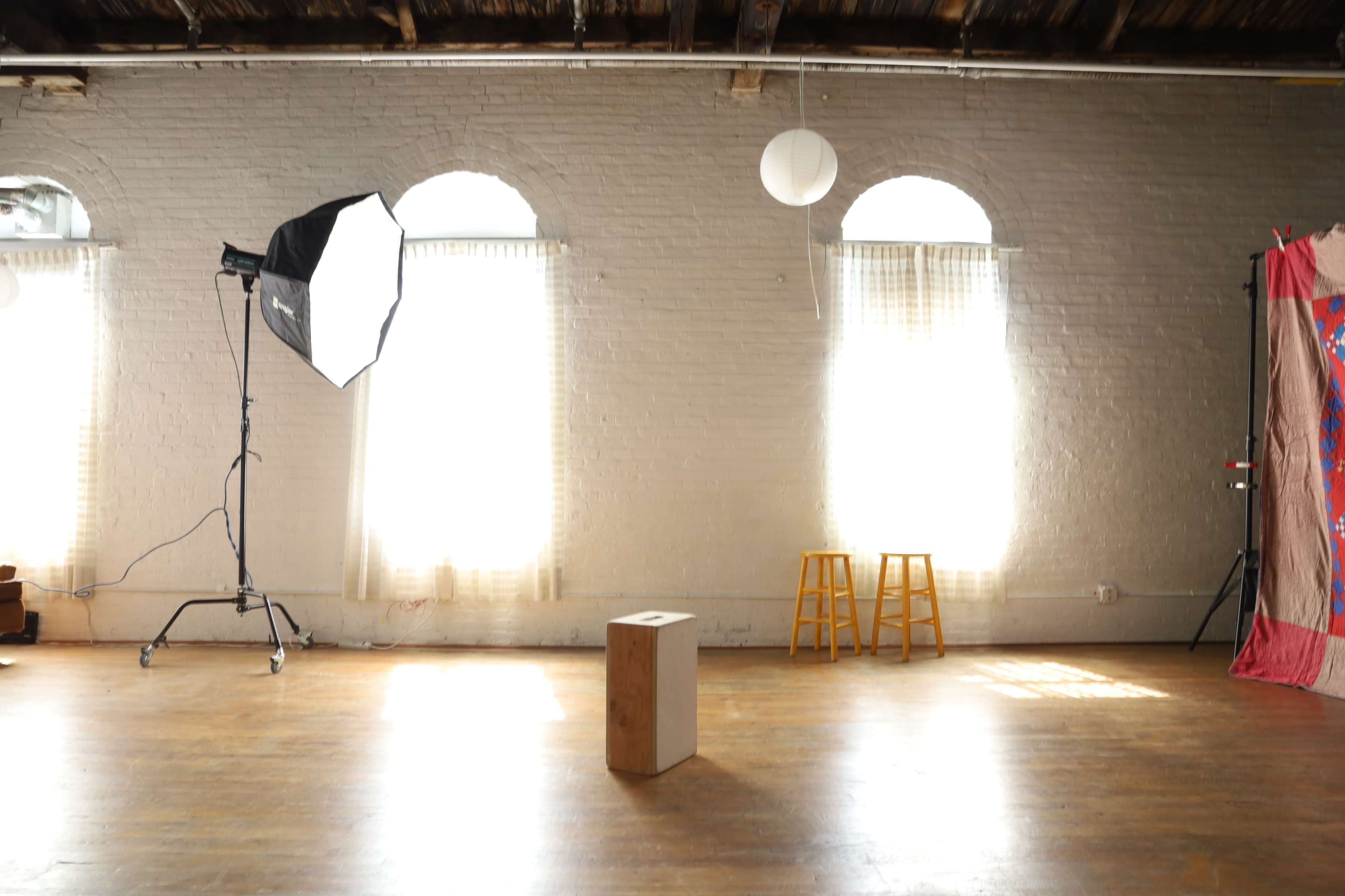 The image shows a well-lit studio space with a wooden box in the center, a photography light setup on the left, and two wooden stools against a wall with windows.