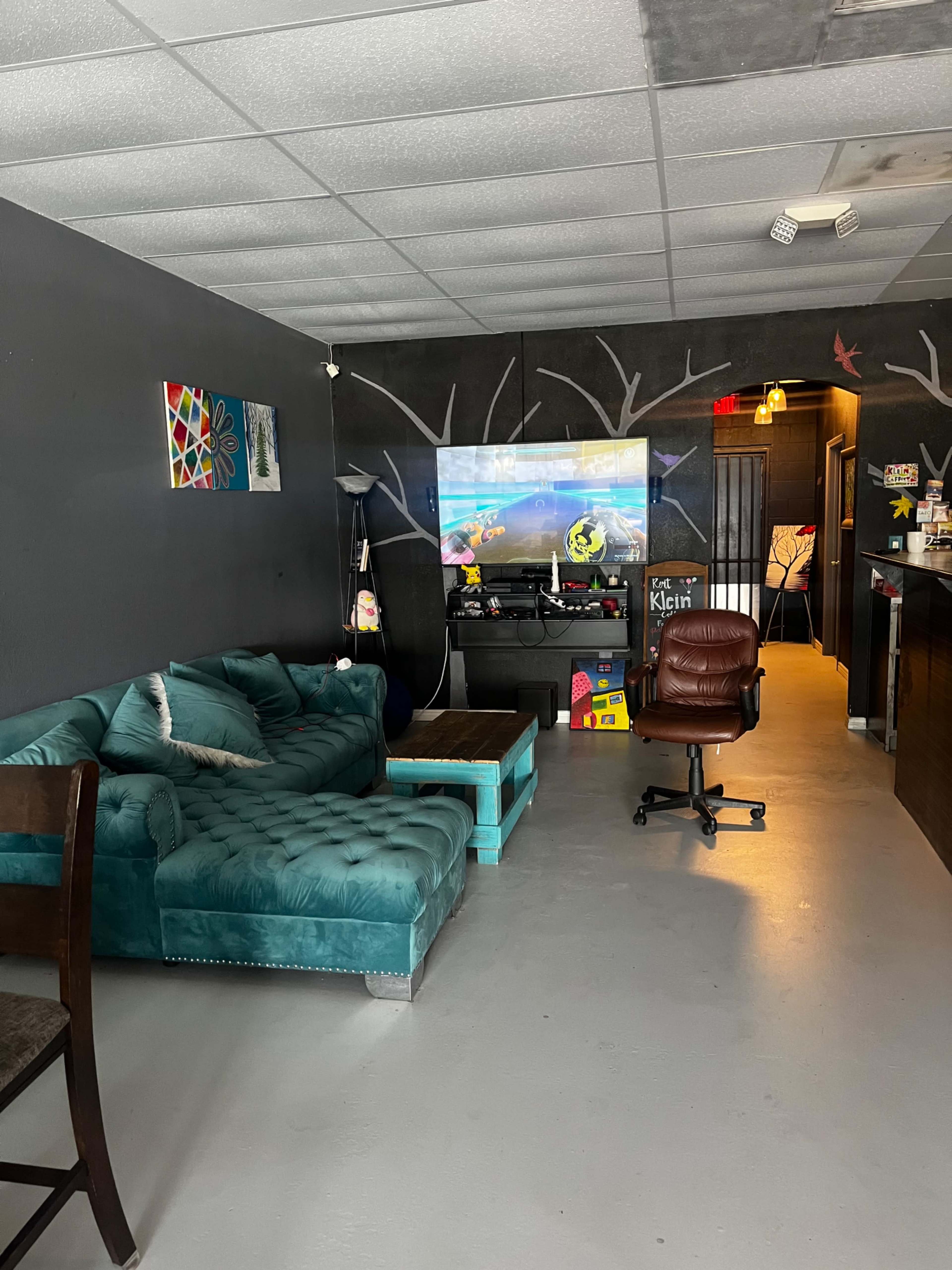 A teal sectional sofa and a wooden coffee table are positioned in a room with gray walls, a television displaying a video game, and an office chair near the entrance.