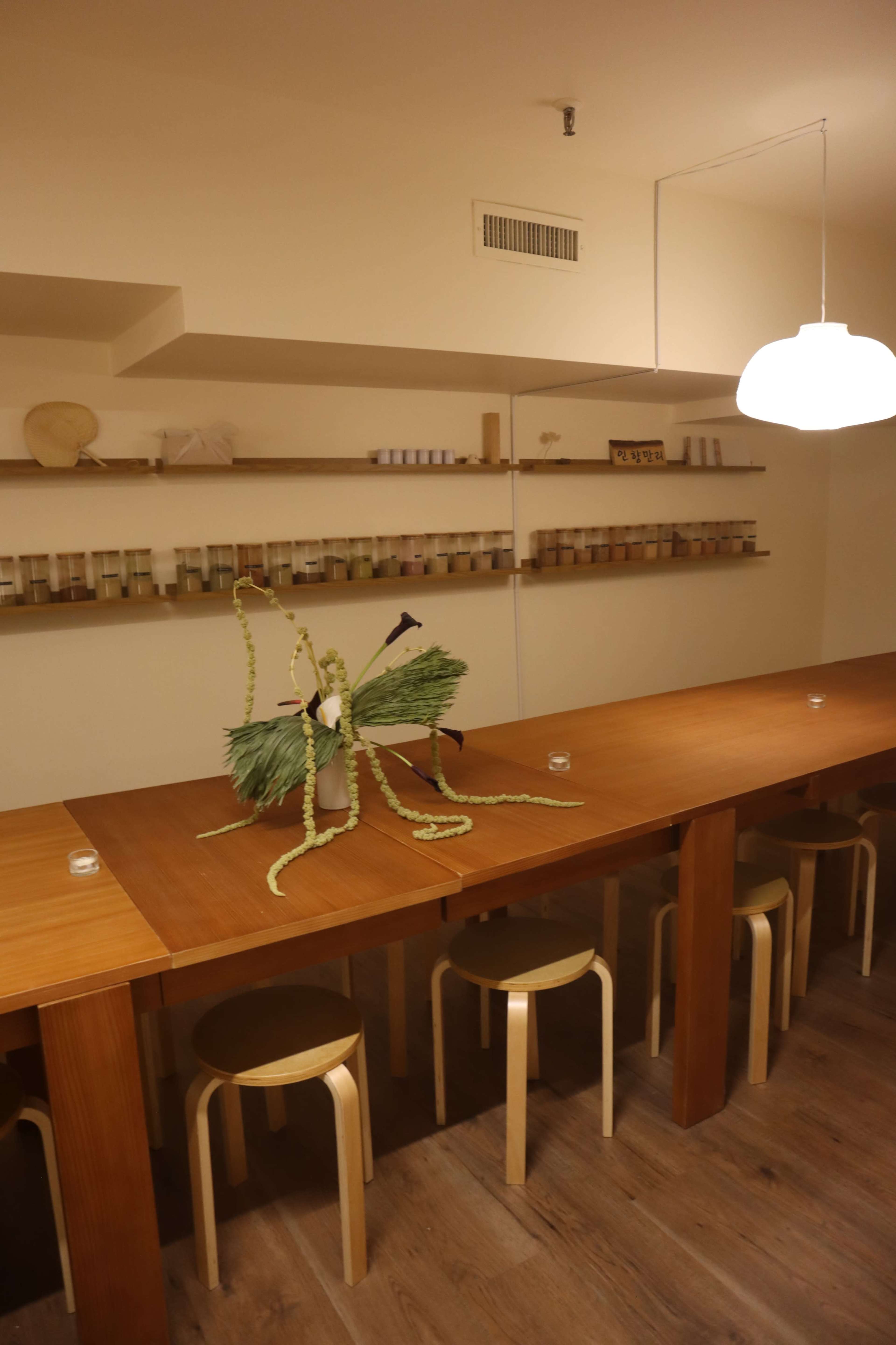 The image shows a minimalist dining space with a long wooden table flanked by several stools, featuring a decorative arrangement of greenery in the center and a shelf with jars in the background.