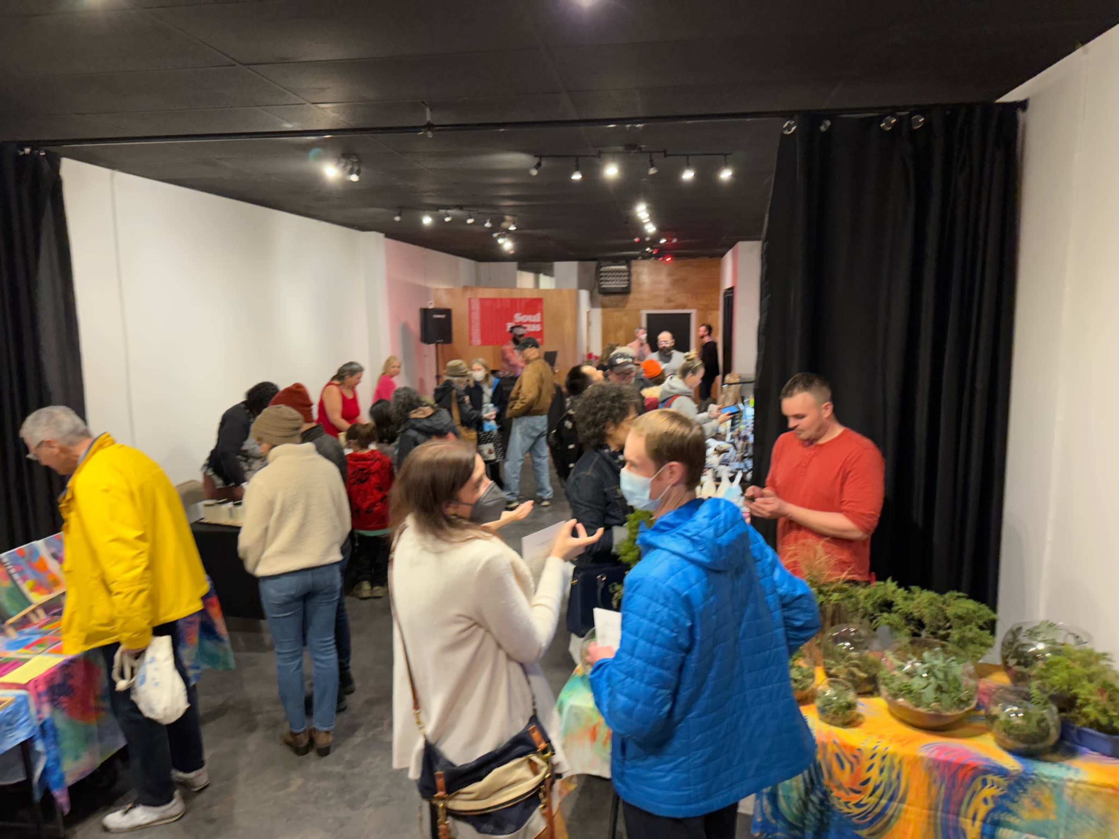 A group of people gathers in a spacious indoor area filled with tables, plants, and decorations, with some engaging in conversations and activities while a small stage is visible in the background.