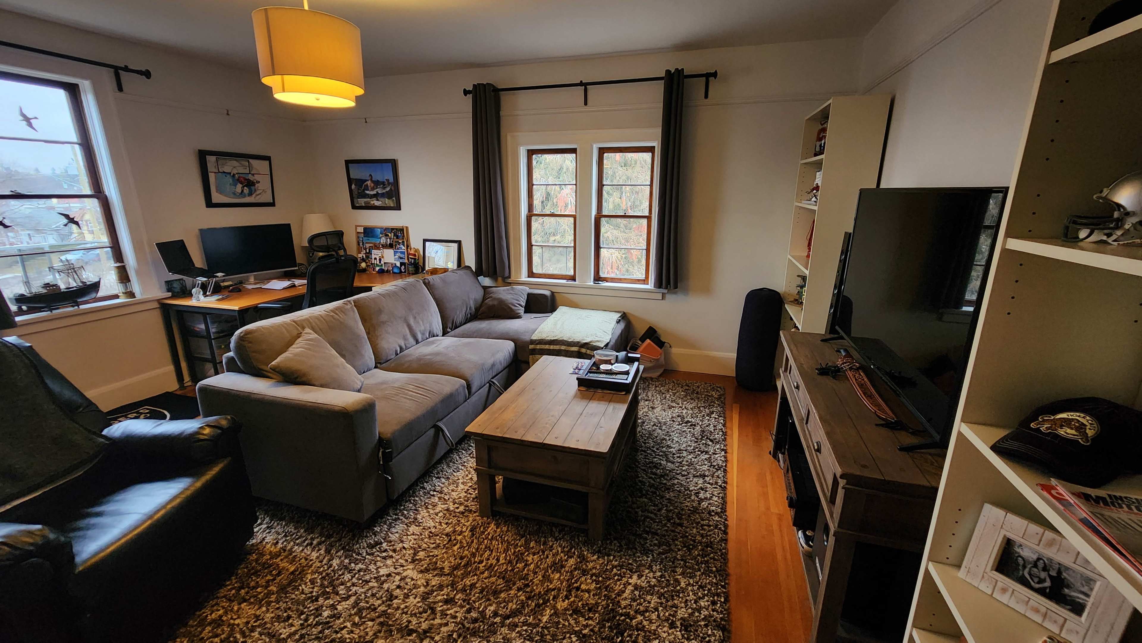 The image shows a living room with a sectional sofa, a coffee table, a television, and two workspaces with computer setups against the walls.