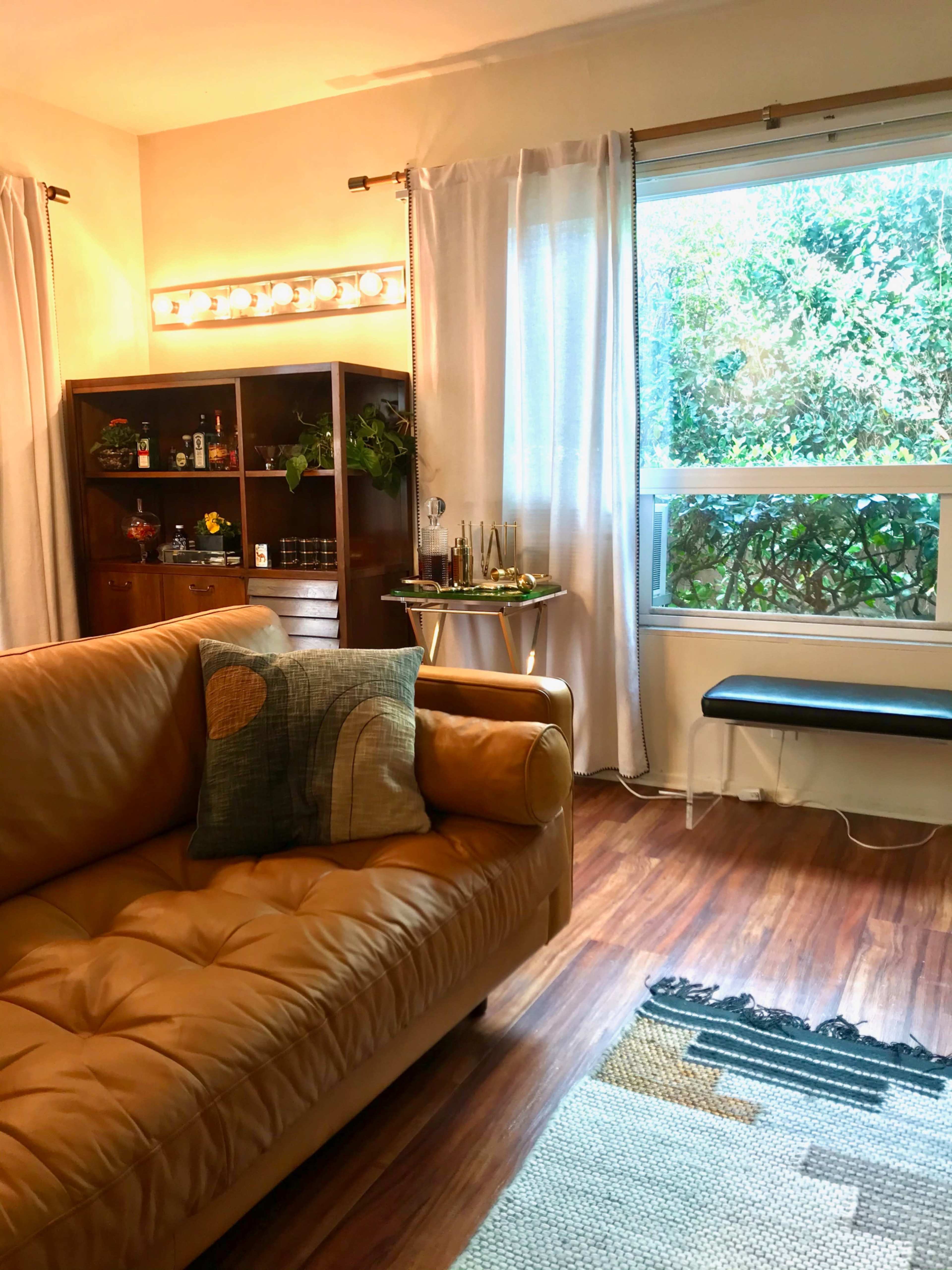 The image shows a furnished living room featuring a brown leather sofa, a wooden shelf with various items, a small table near a window, and a patterned rug on the floor.