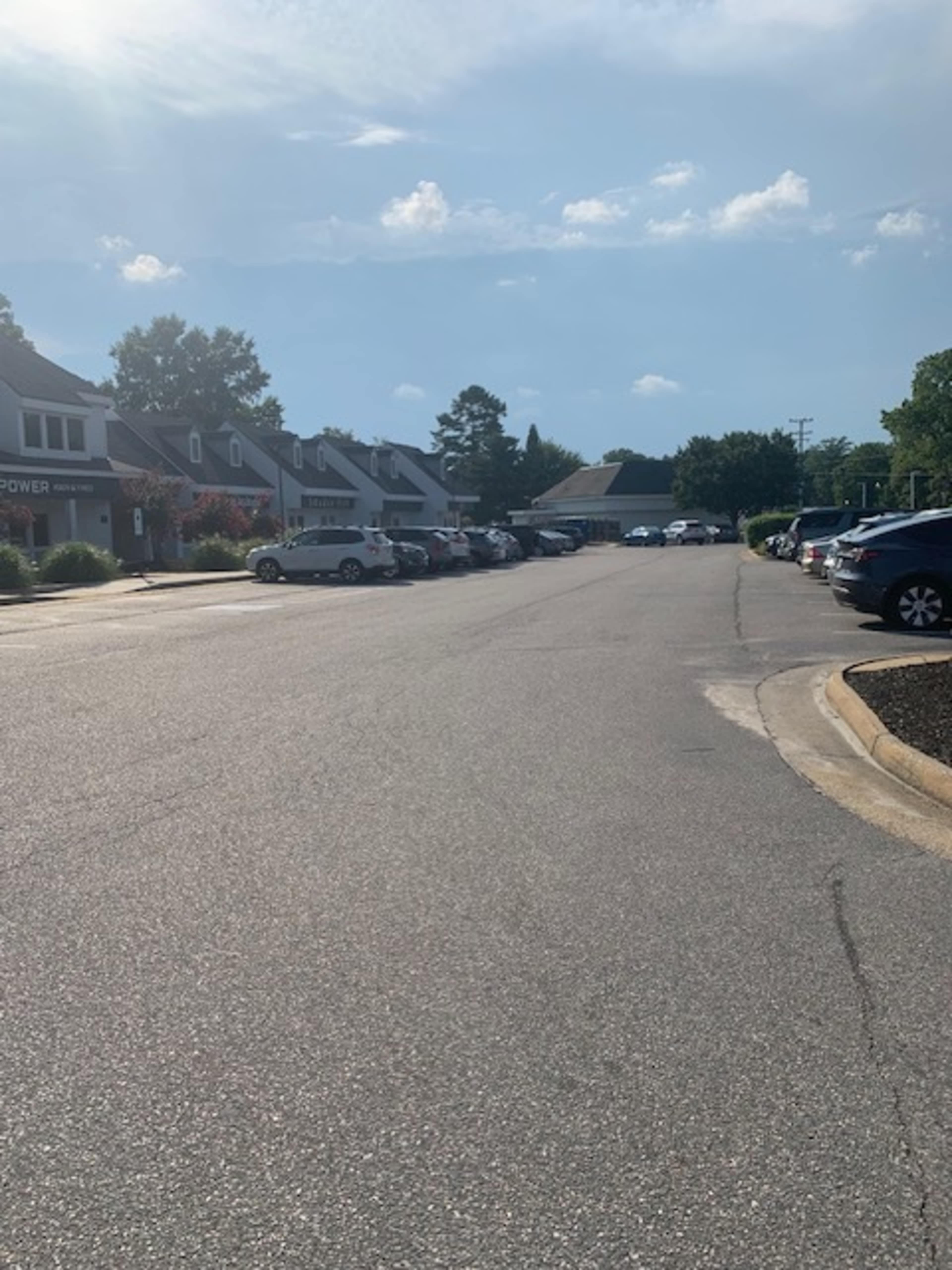 A street lined with parked cars along both sides, with buildings and trees in the background under a clear blue sky.