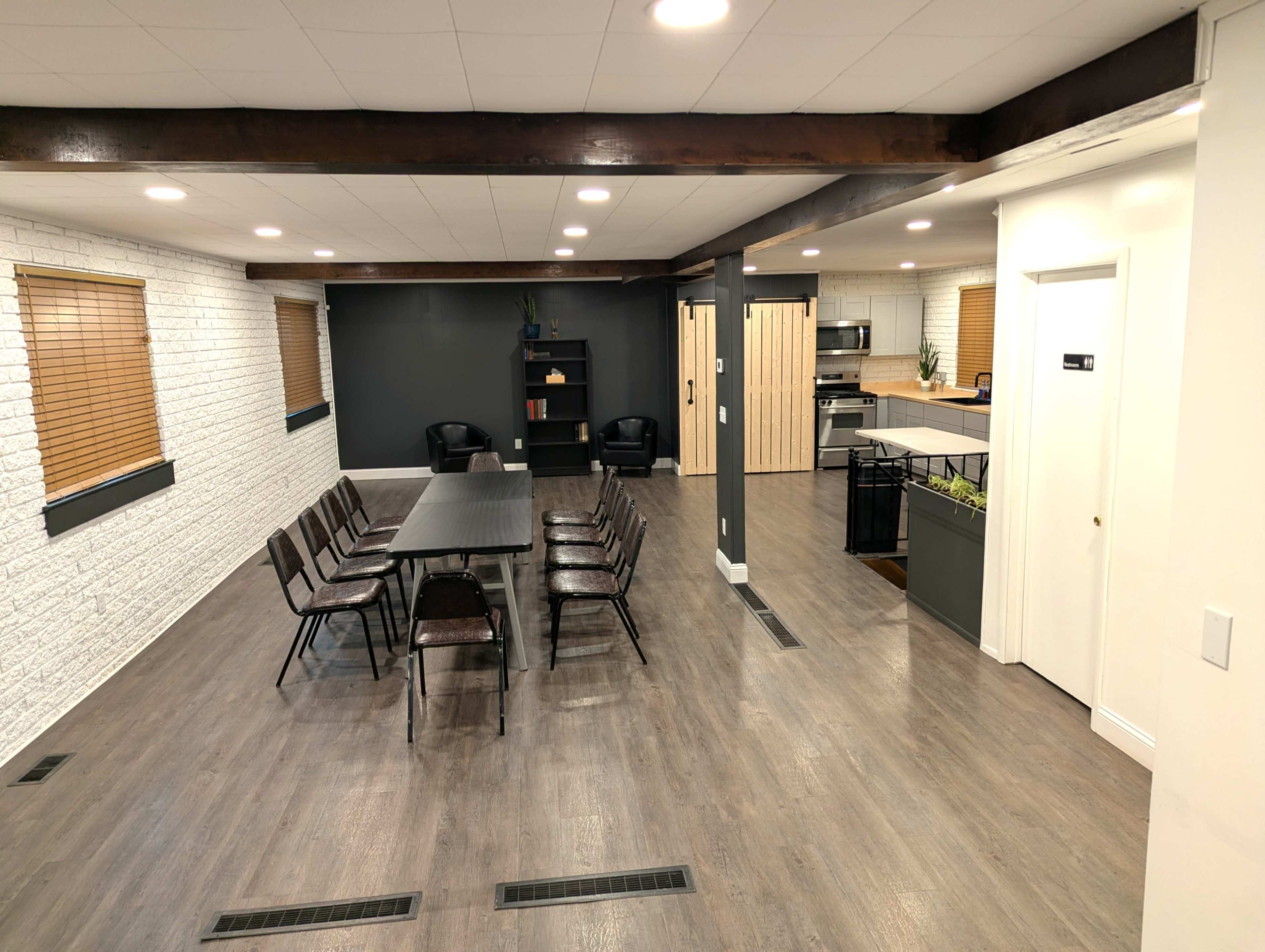 The image shows a modern, open-concept room with a large dining table surrounded by chairs, a kitchenette in the background, and a mix of light and dark wall colors.