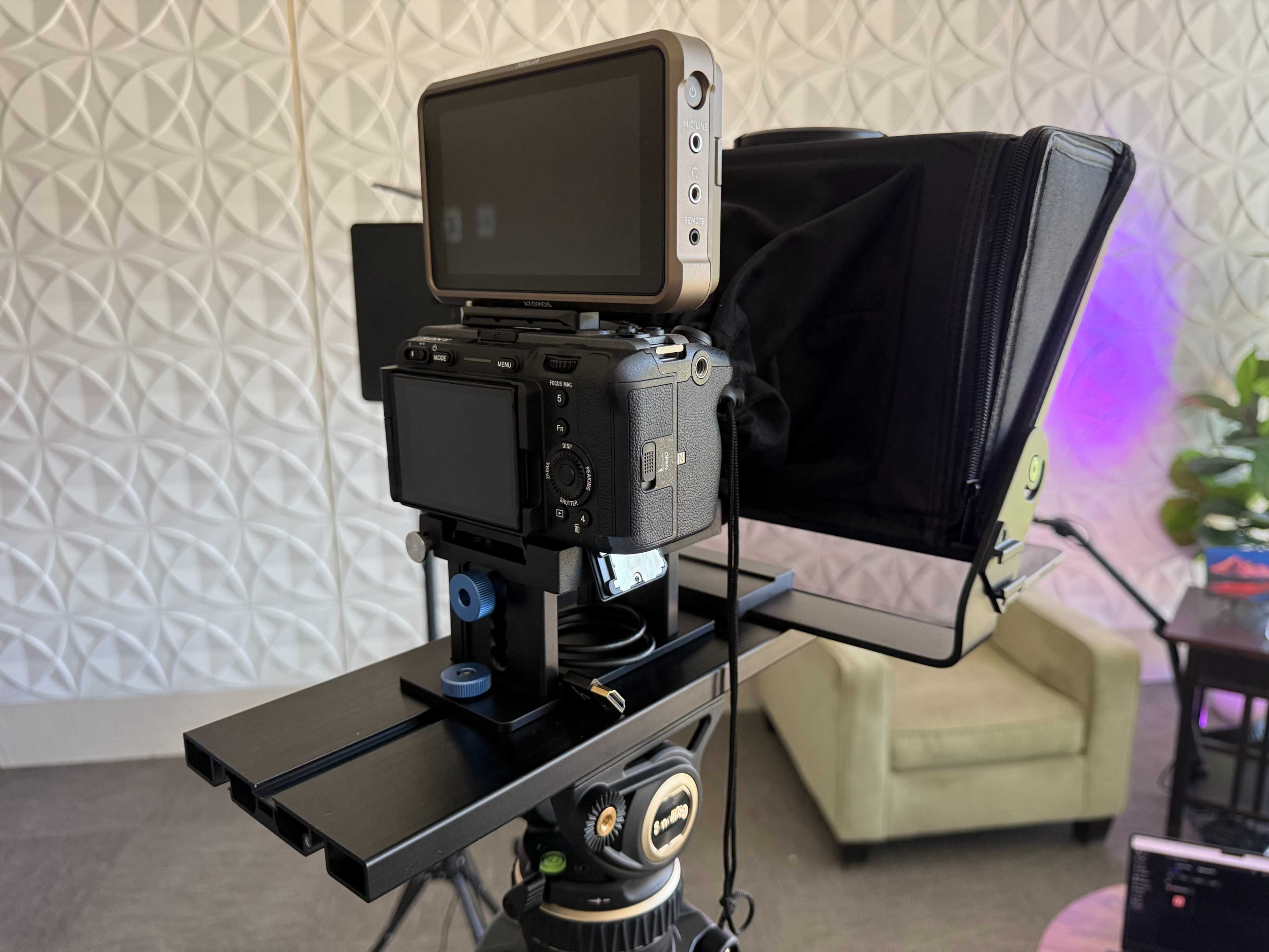 A camera setup is mounted on a tripod, featuring a monitor and a lens hood in a well-lit room.
