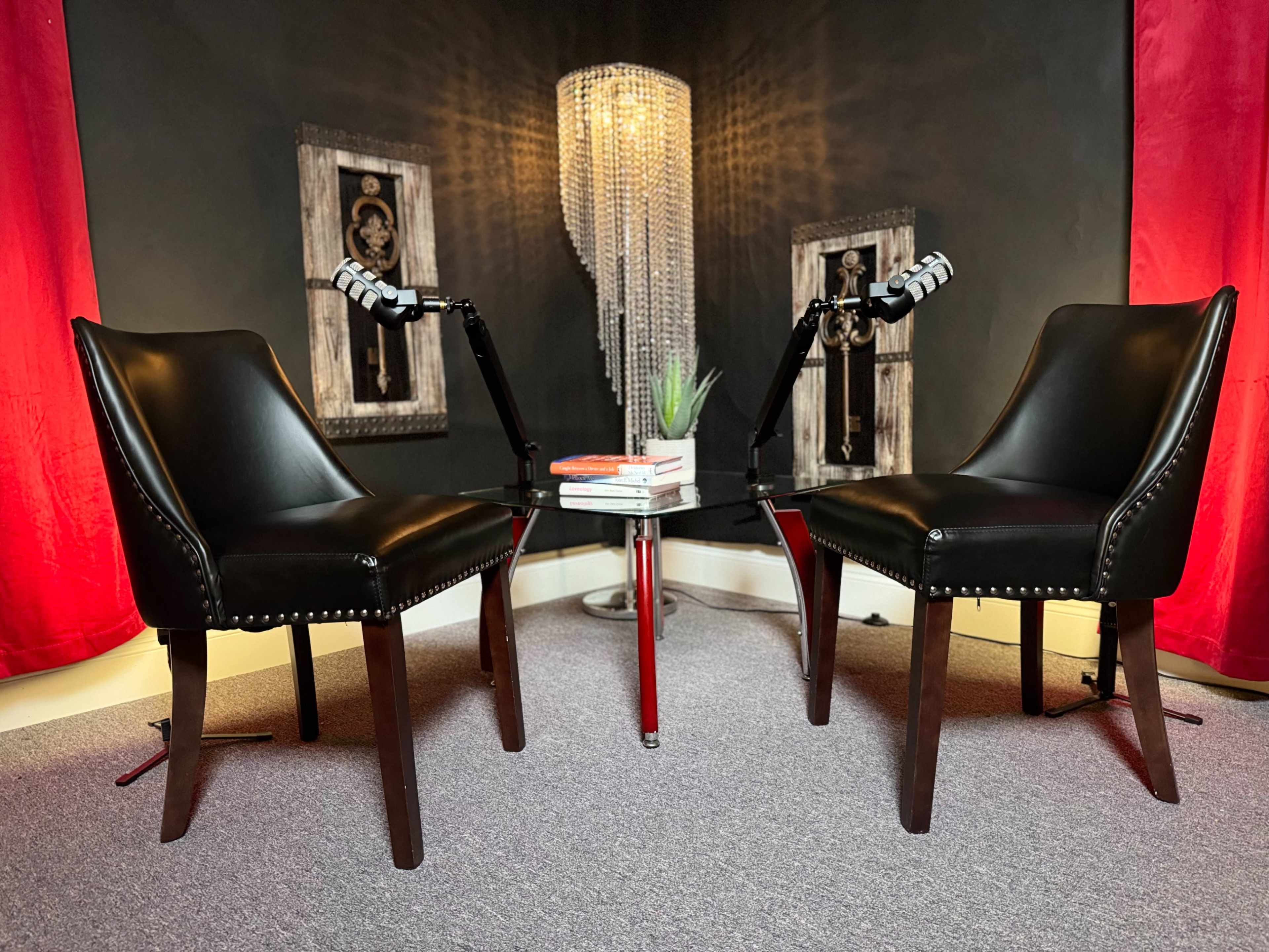 The image features two black leather chairs facing each other with a glass table in between, under a chandelier, against a backdrop of red curtains and wall art.