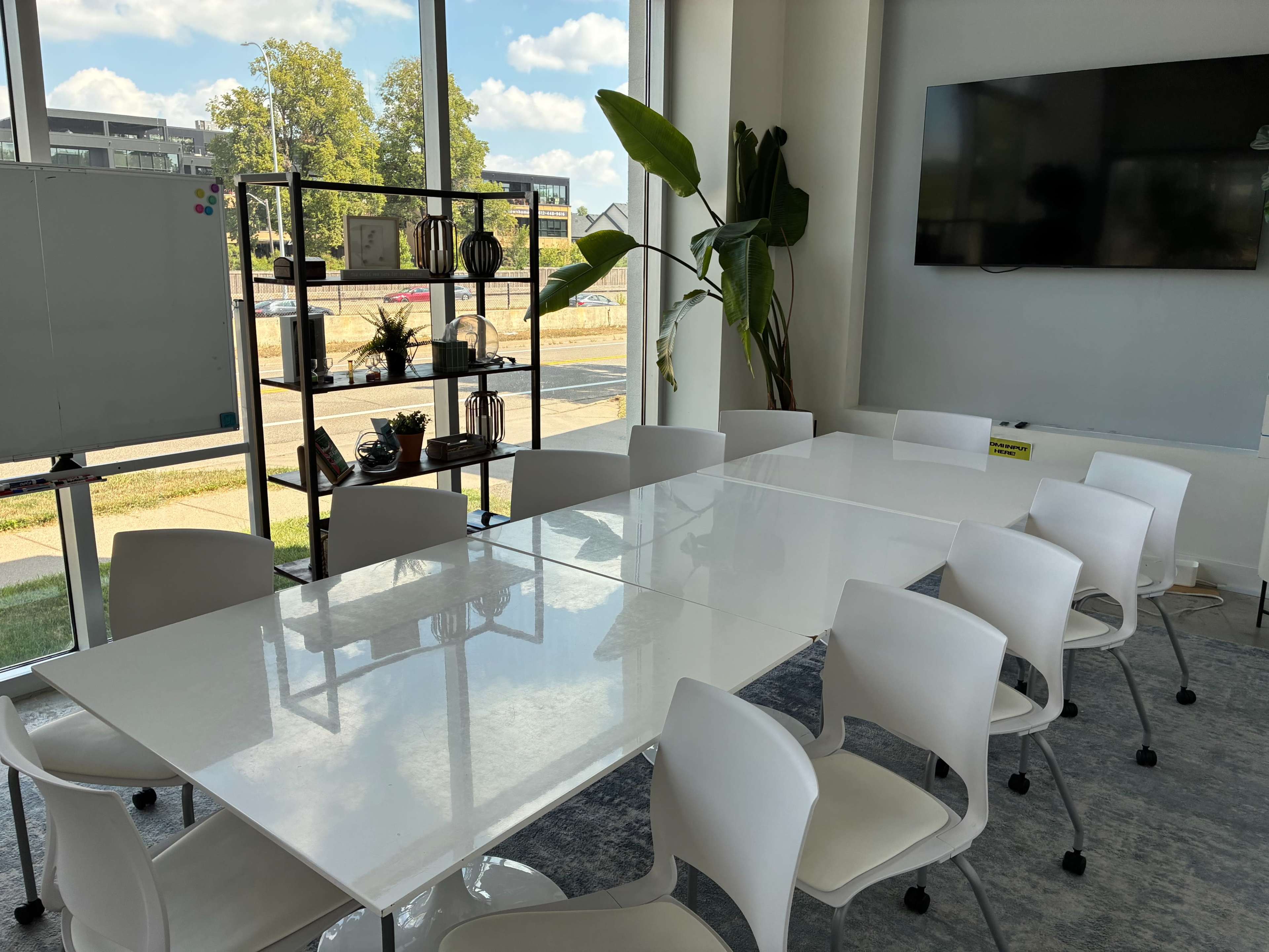 A bright conference room features a large white table surrounded by chairs, with a TV screen on the wall and a window showcasing an outdoor view.