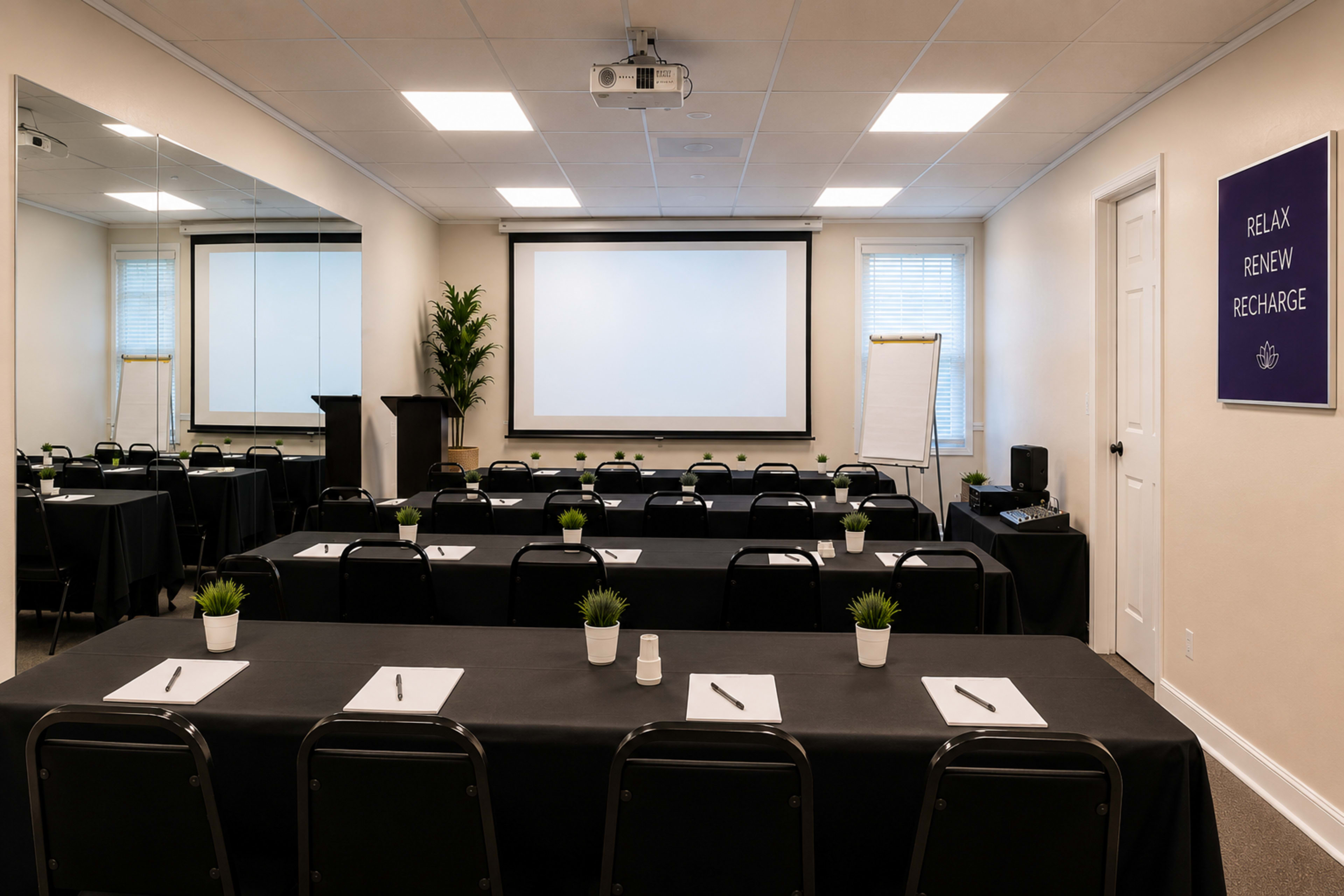 The image shows a conference room set up with rows of black tables and chairs, a projector screen at the front, and plants on the tables.