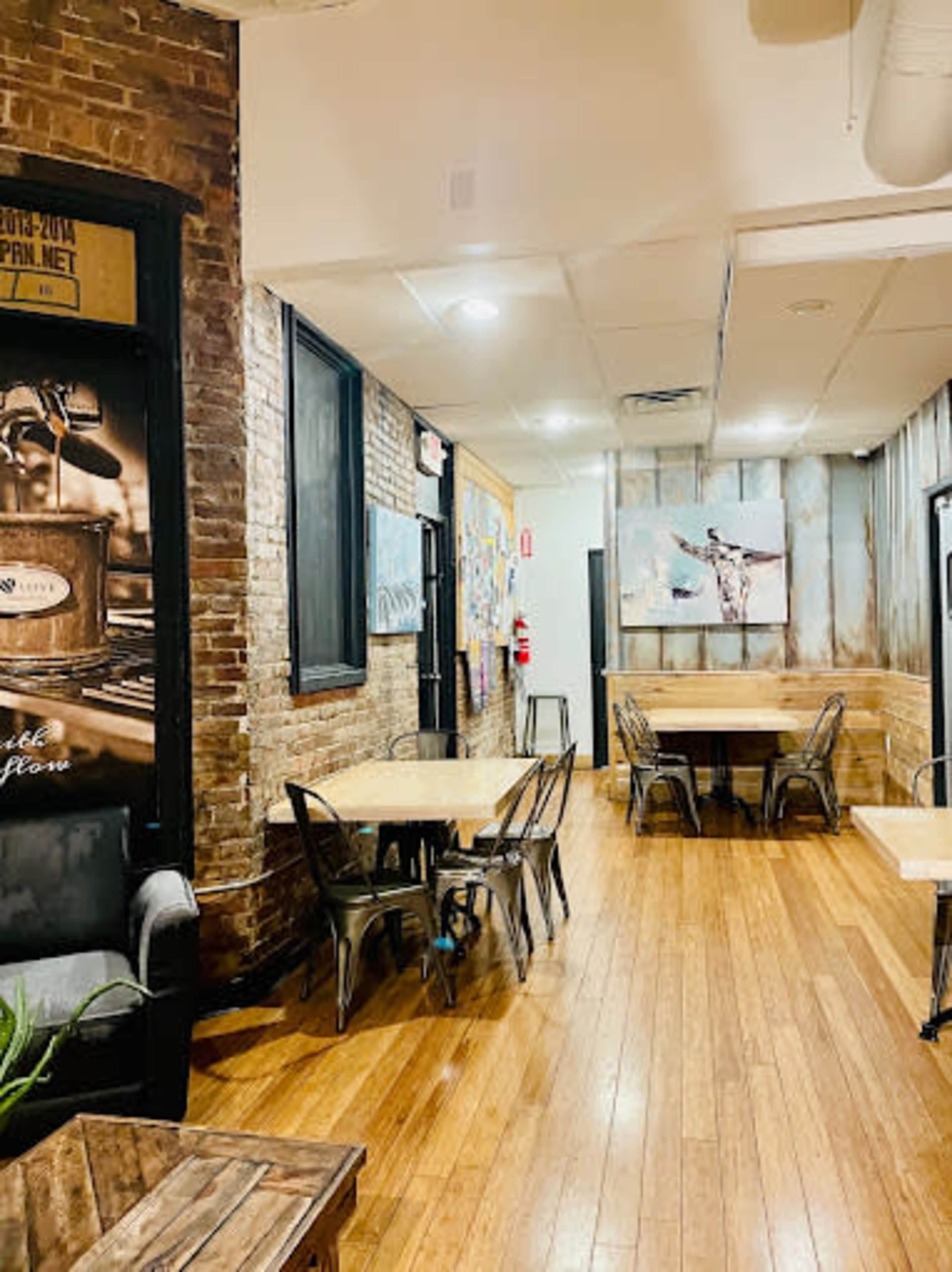 The image shows a cozy café interior with wooden floors, exposed brick walls, and metal chairs arranged around small tables.