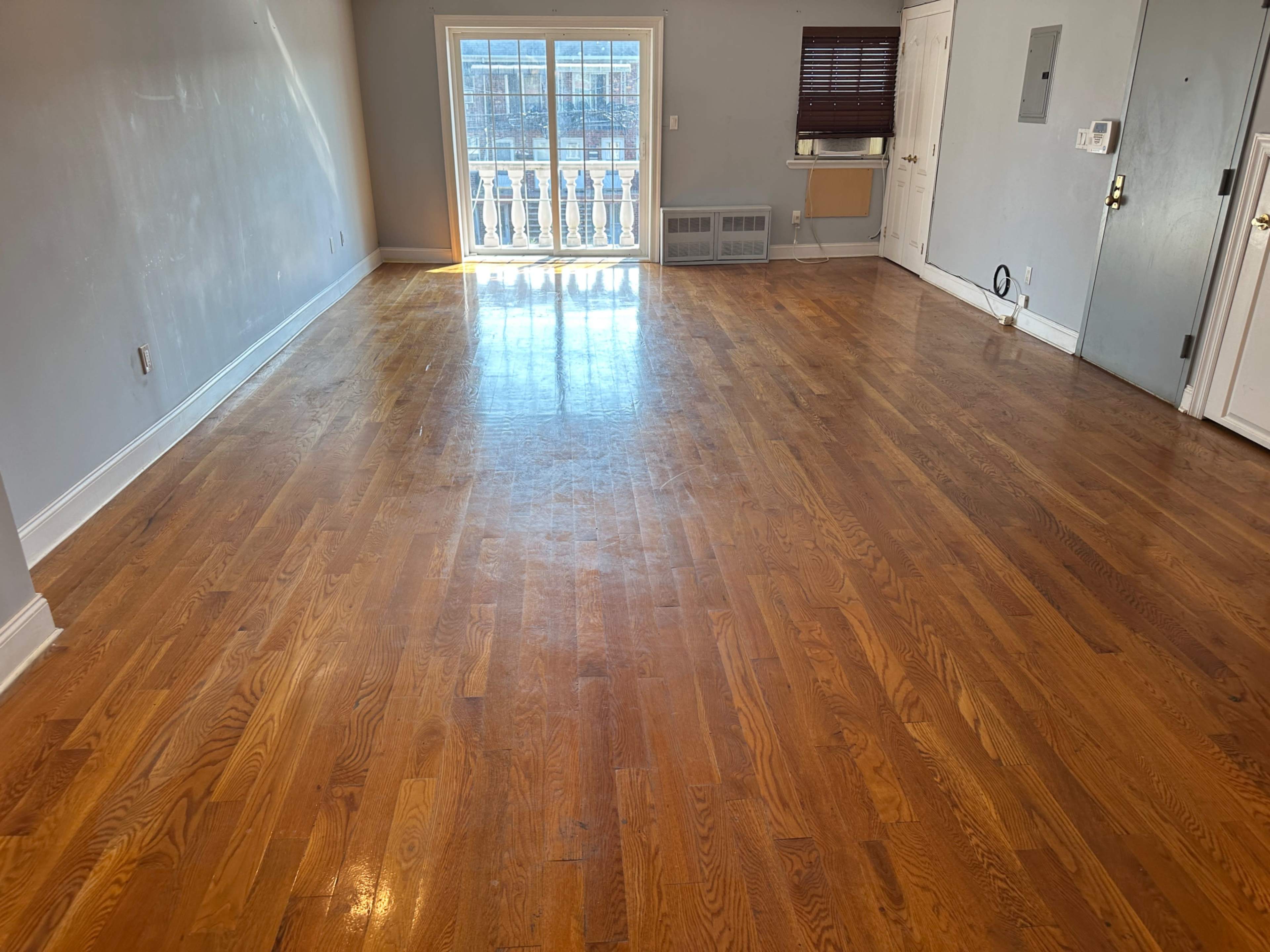 The image shows an empty room with hardwood flooring and a large window that lets in natural light.