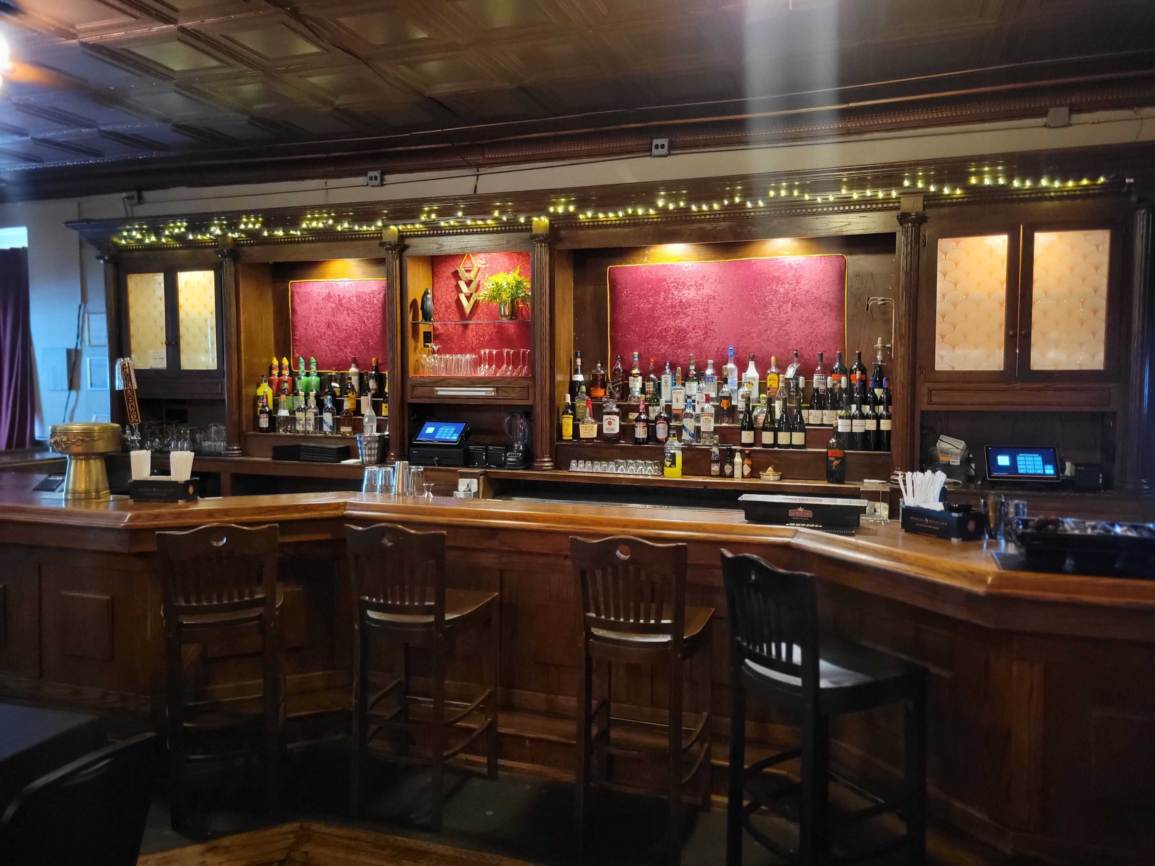 The image shows a wooden bar with shelves displaying a variety of bottles and a row of chairs for patrons.