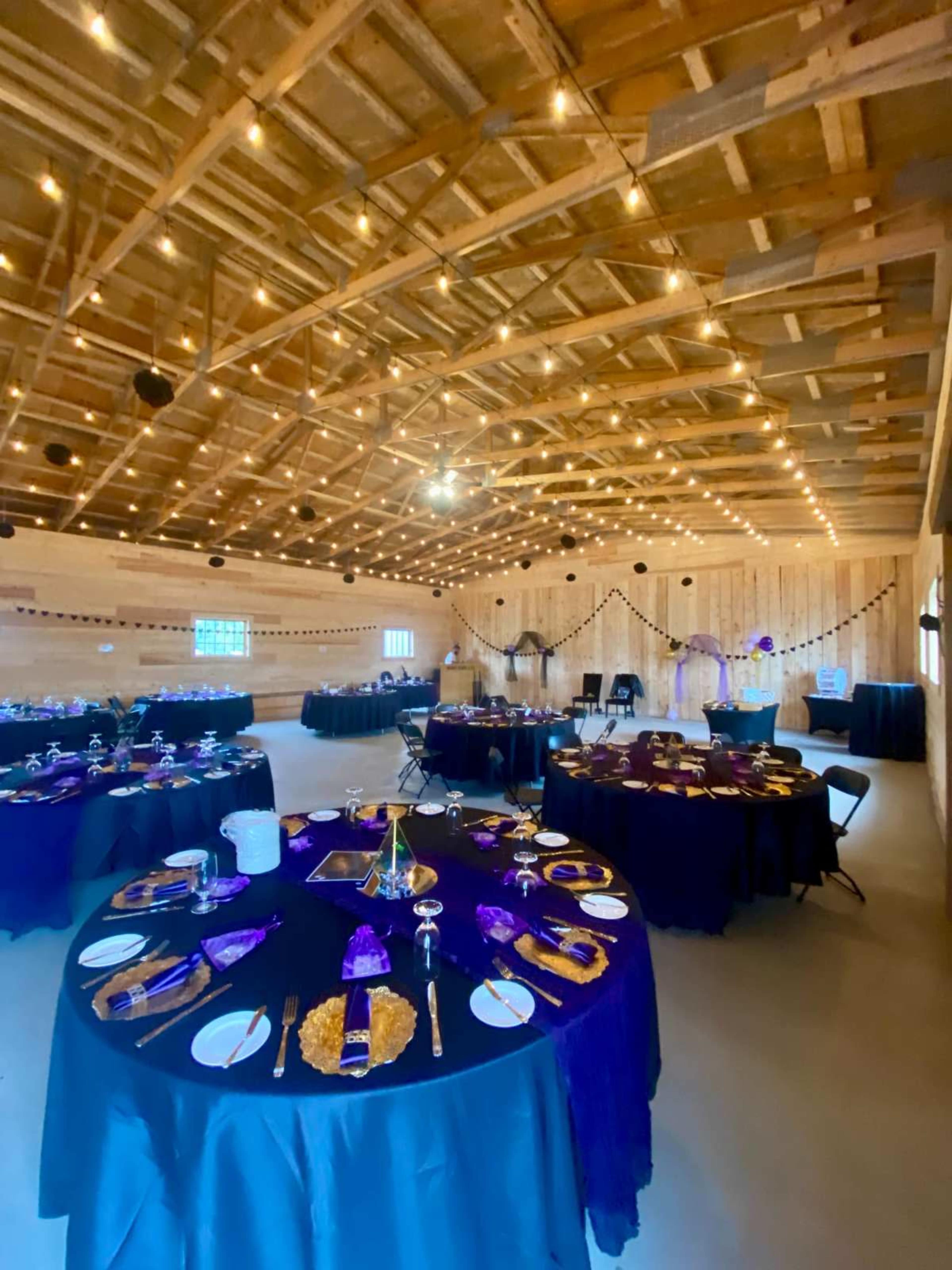 The image shows a decorated barn interior with several round tables set for an event, featuring black tablecloths and purple accents.