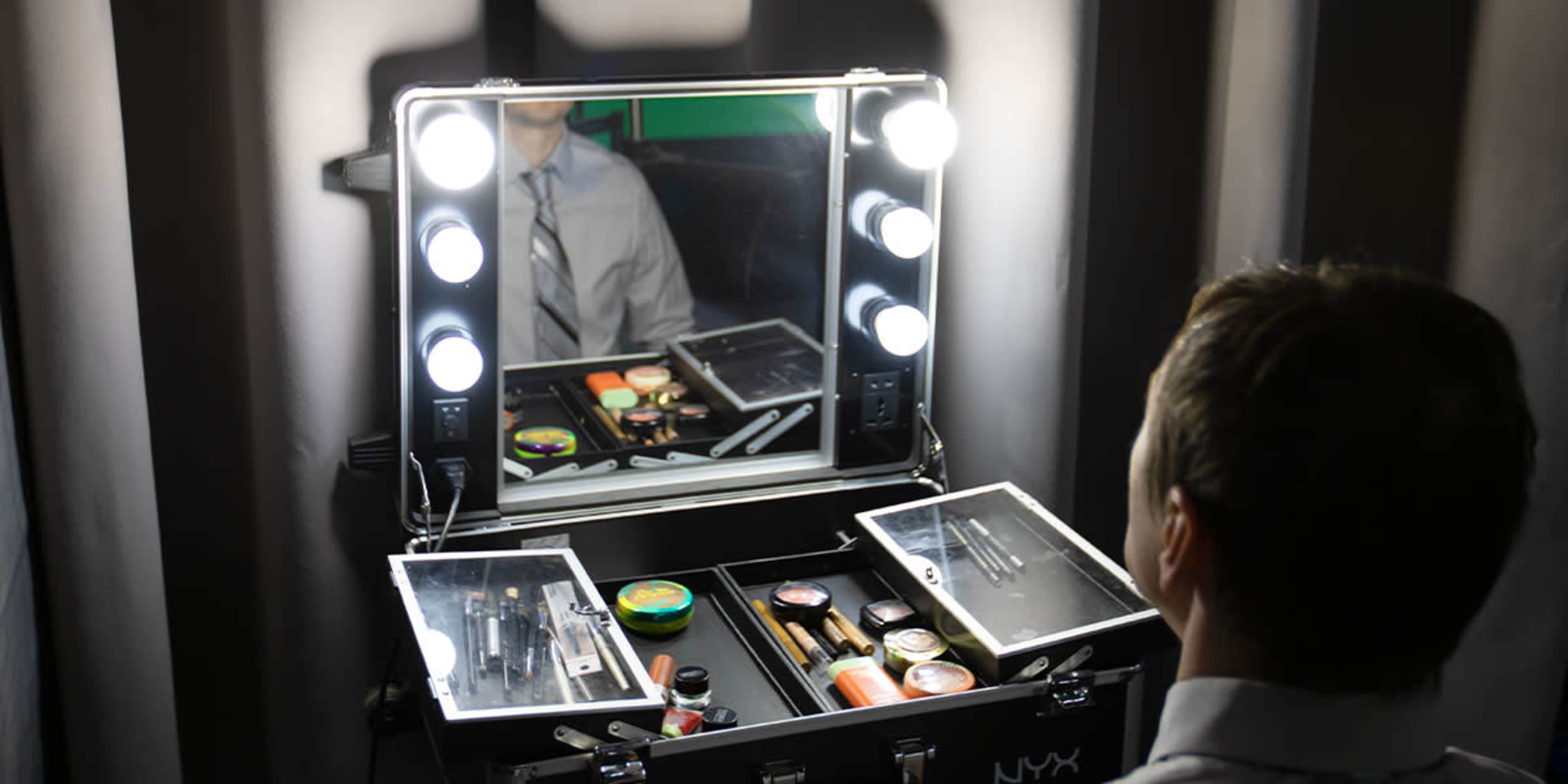 A person in a tie is sitting in front of a well-lit makeup mirror with various cosmetics arranged in an open case.