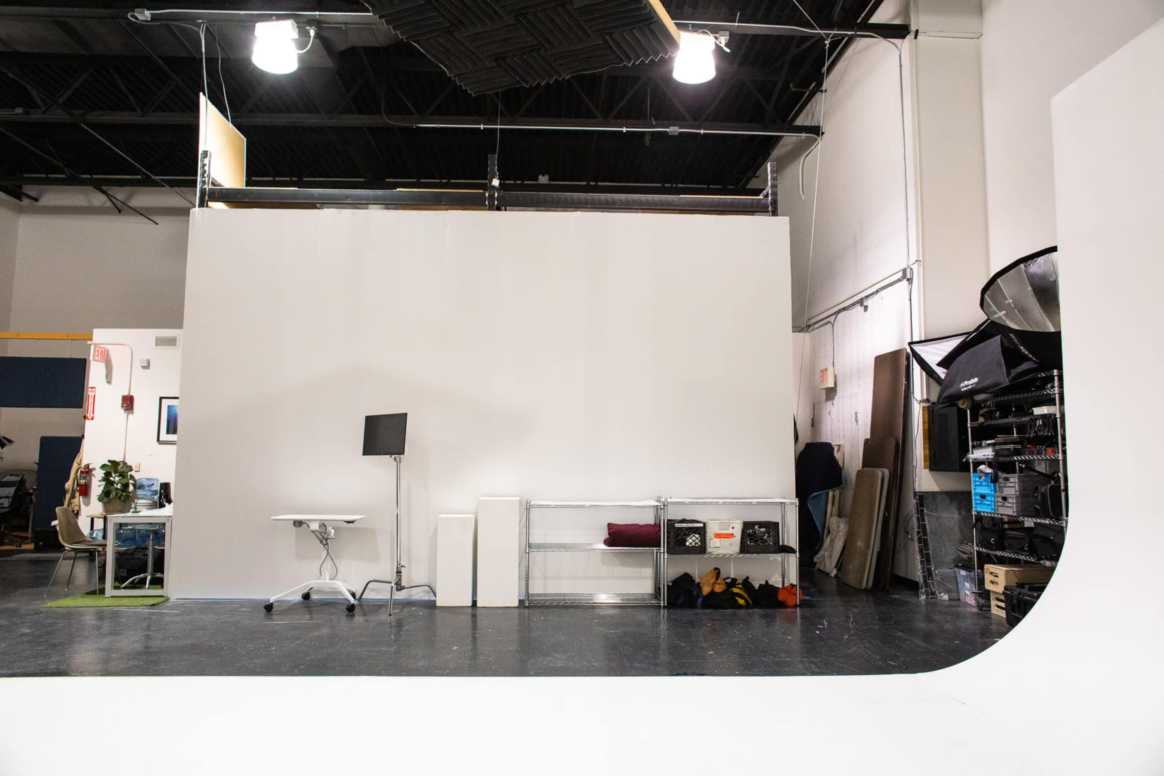 A spacious photography studio with a white backdrop, a desk, and various equipment along the side.
