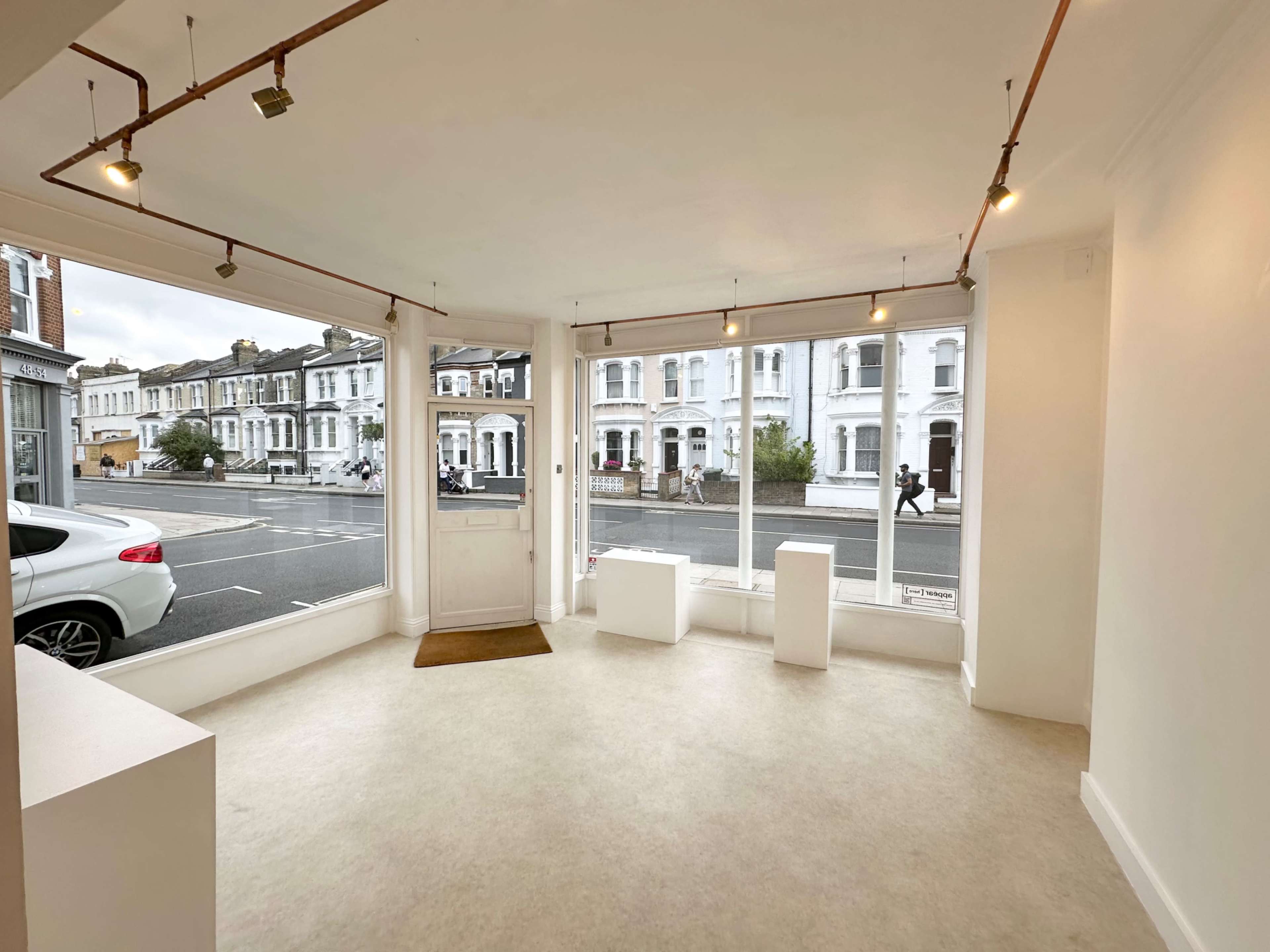 The image shows an empty commercial space with large windows facing a street, featuring minimalistic white walls and a light-colored floor.