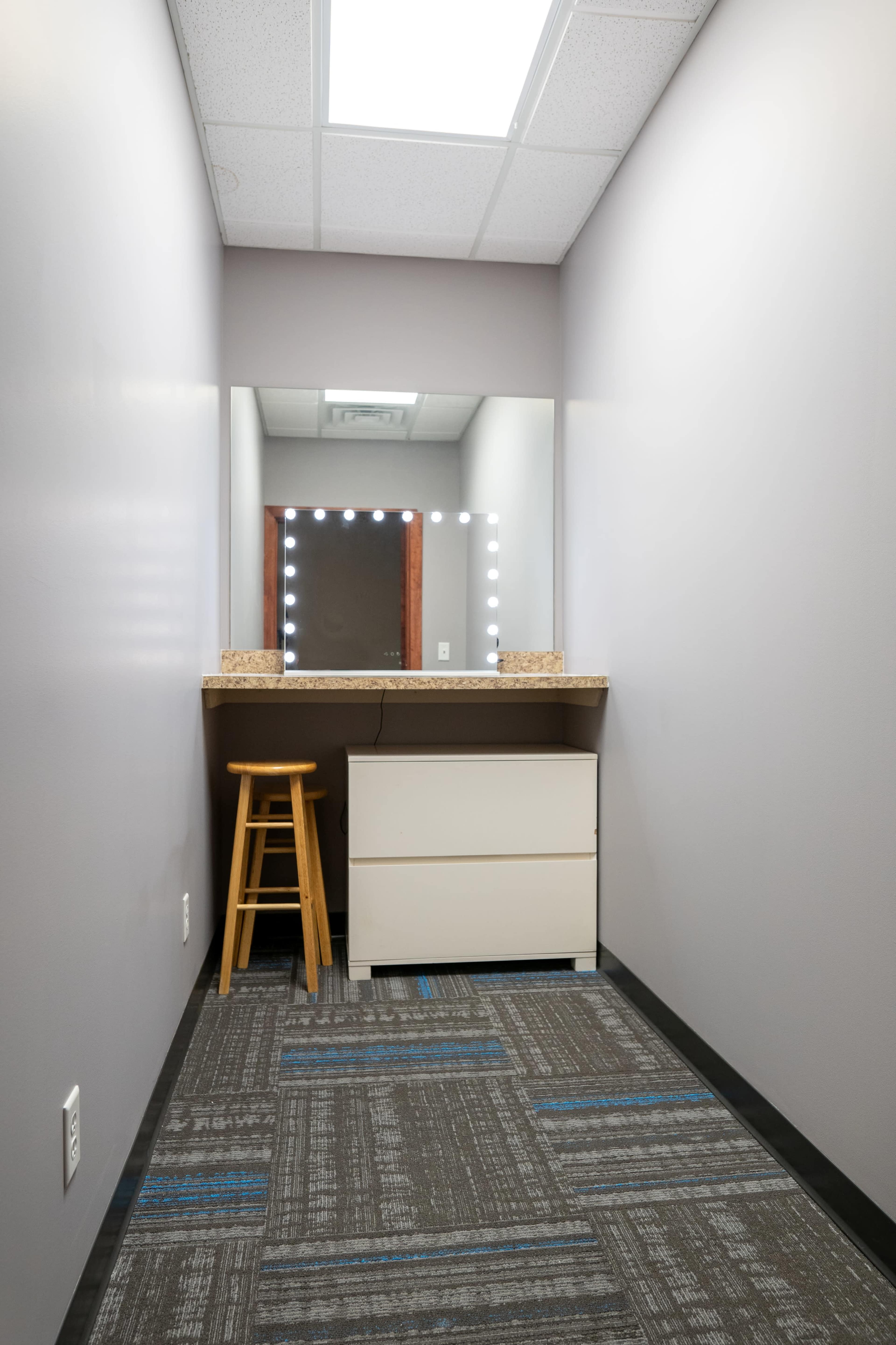 The image shows a narrow, well-lit vanity area with a mirror and a stool against a plain wall, featuring a drawer unit below the countertop.