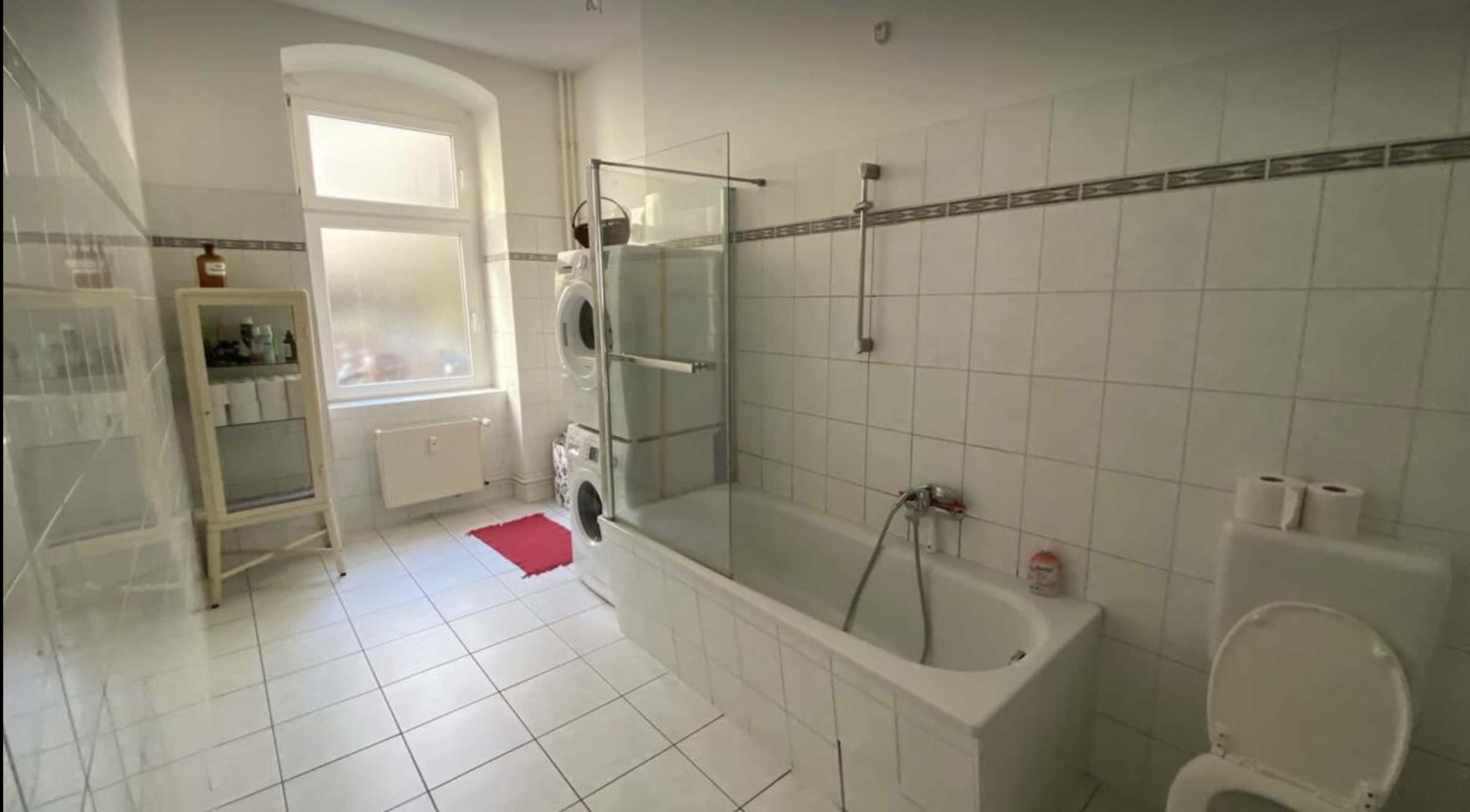 A bright bathroom with a bathtub, glass shower partition, washing machine, and a small window allowing natural light.