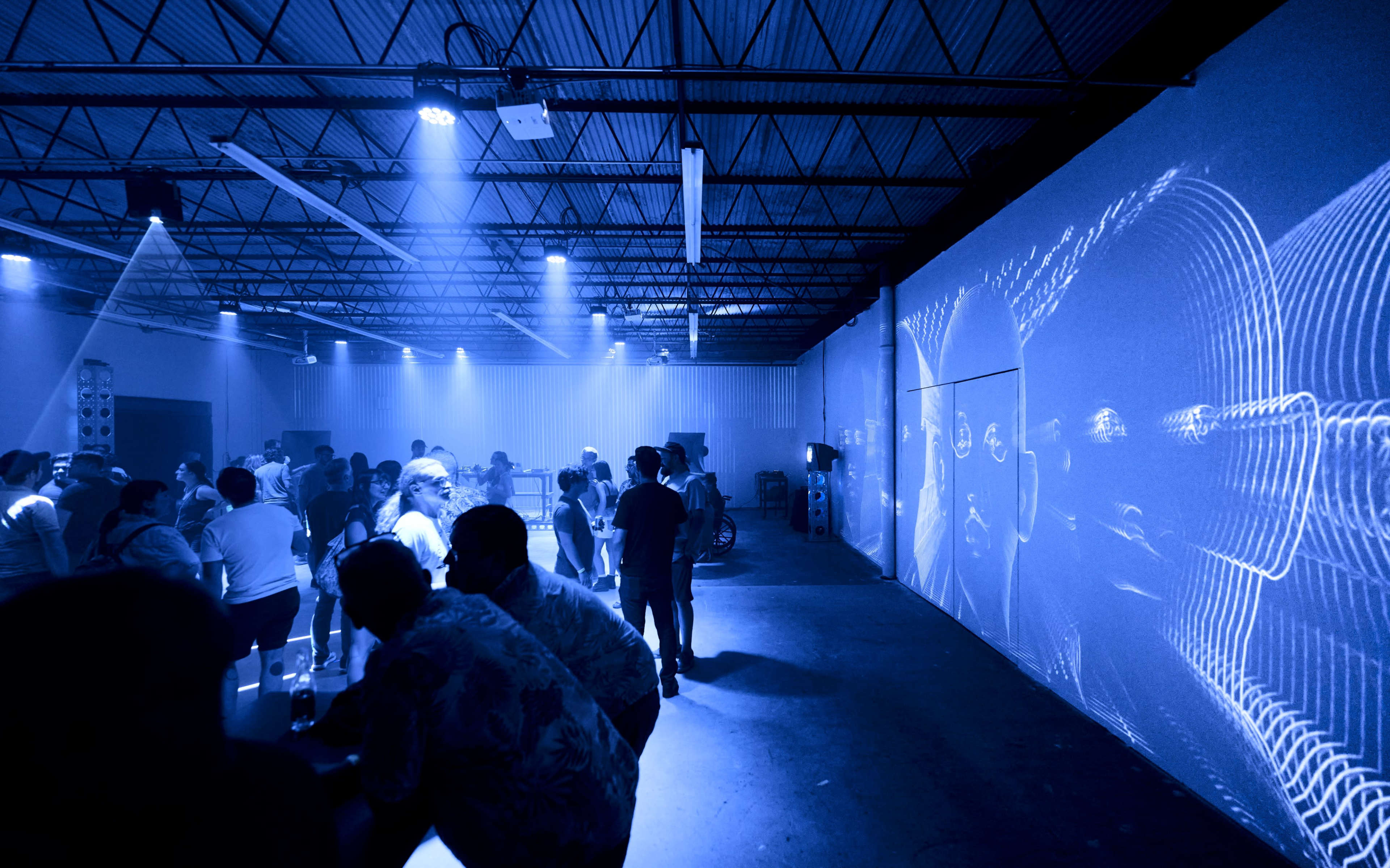 A dimly lit warehouse space features a crowd of people dancing and interacting, with blue light projections on the walls.