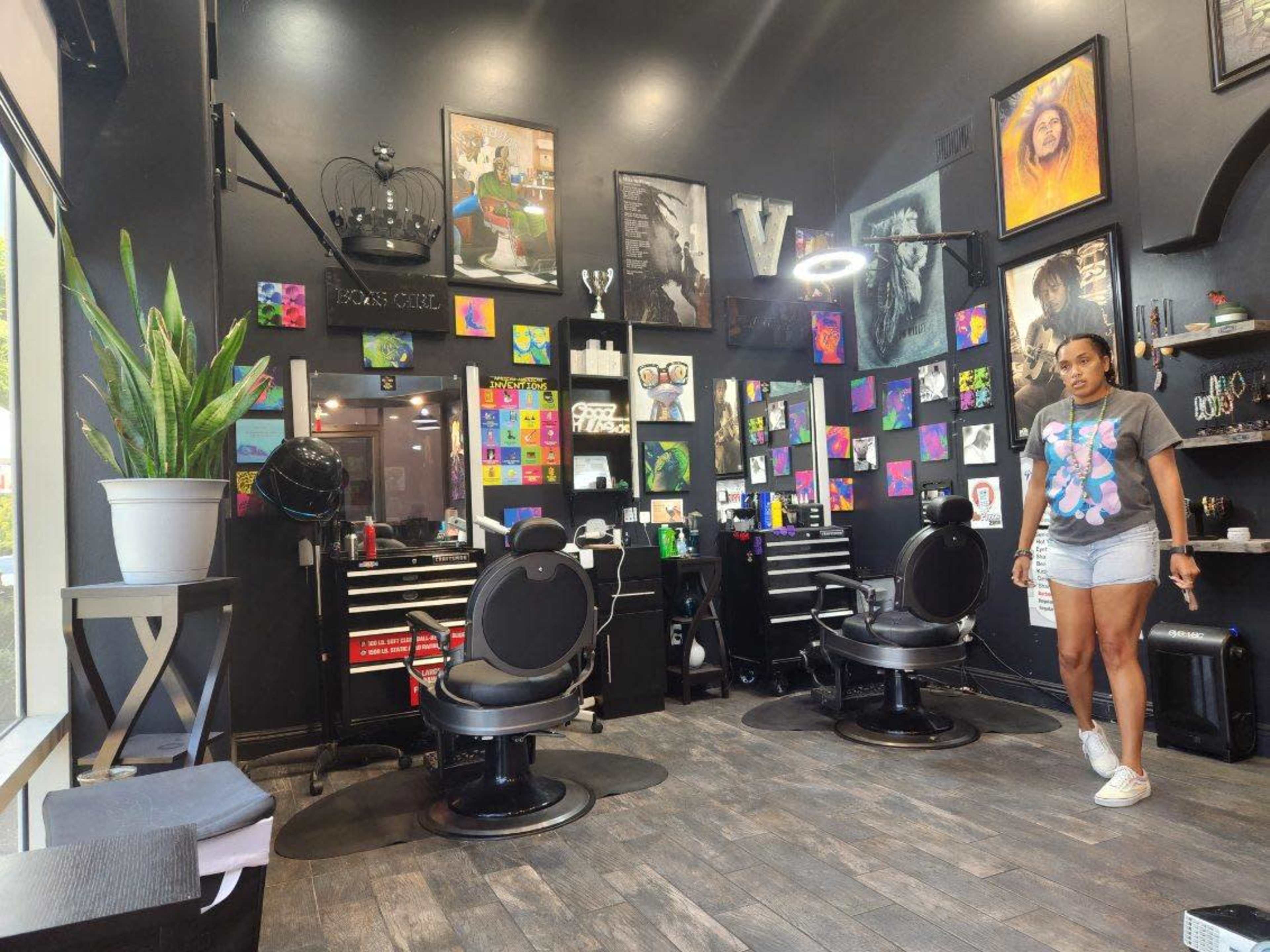The interior of a hair salon features two black styling chairs, a wall adorned with colorful artwork, and a woman walking through the space.