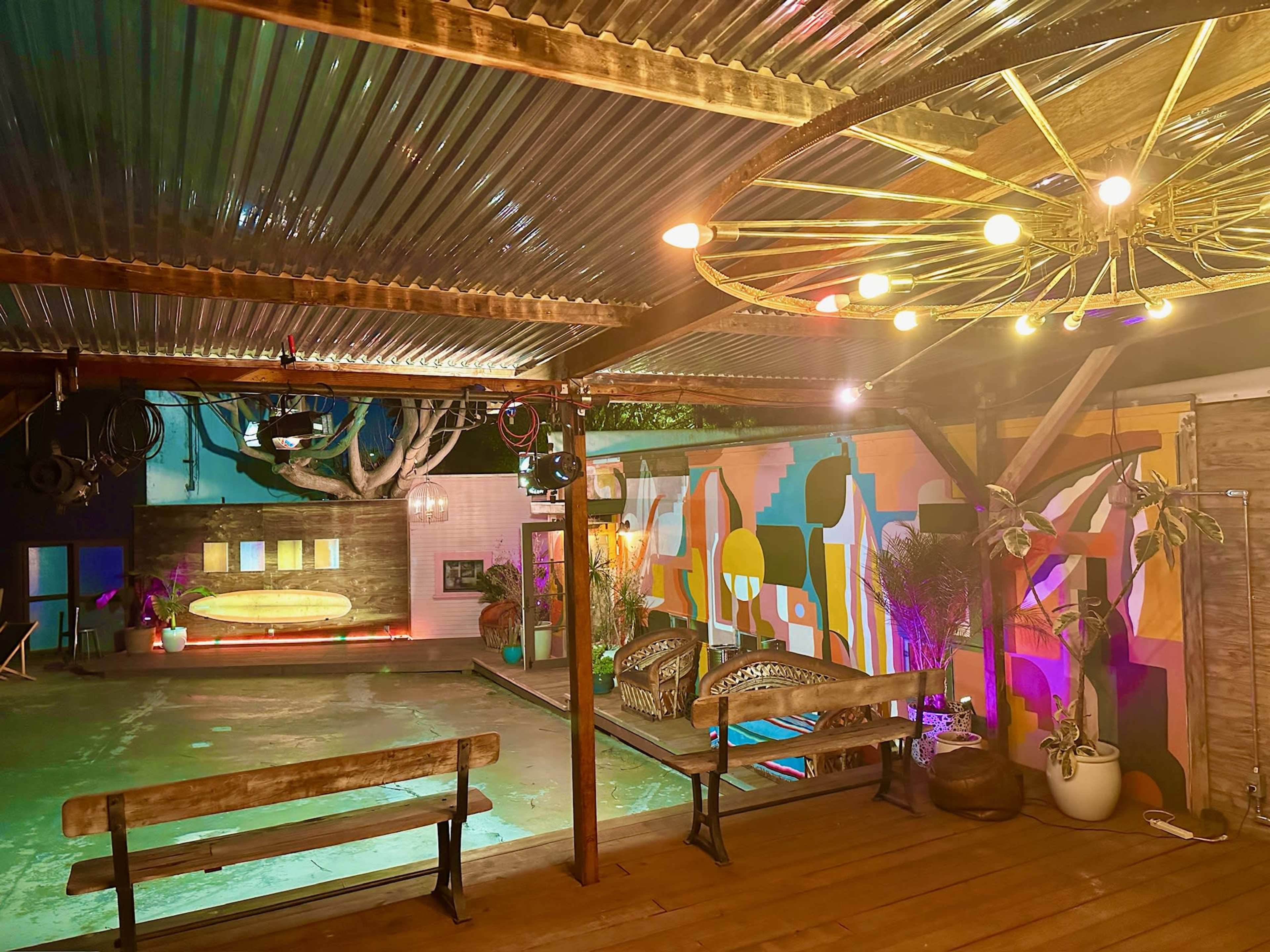 A colorful outdoor space with a pool, wooden benches, and decorative lights hanging from a metal roof.