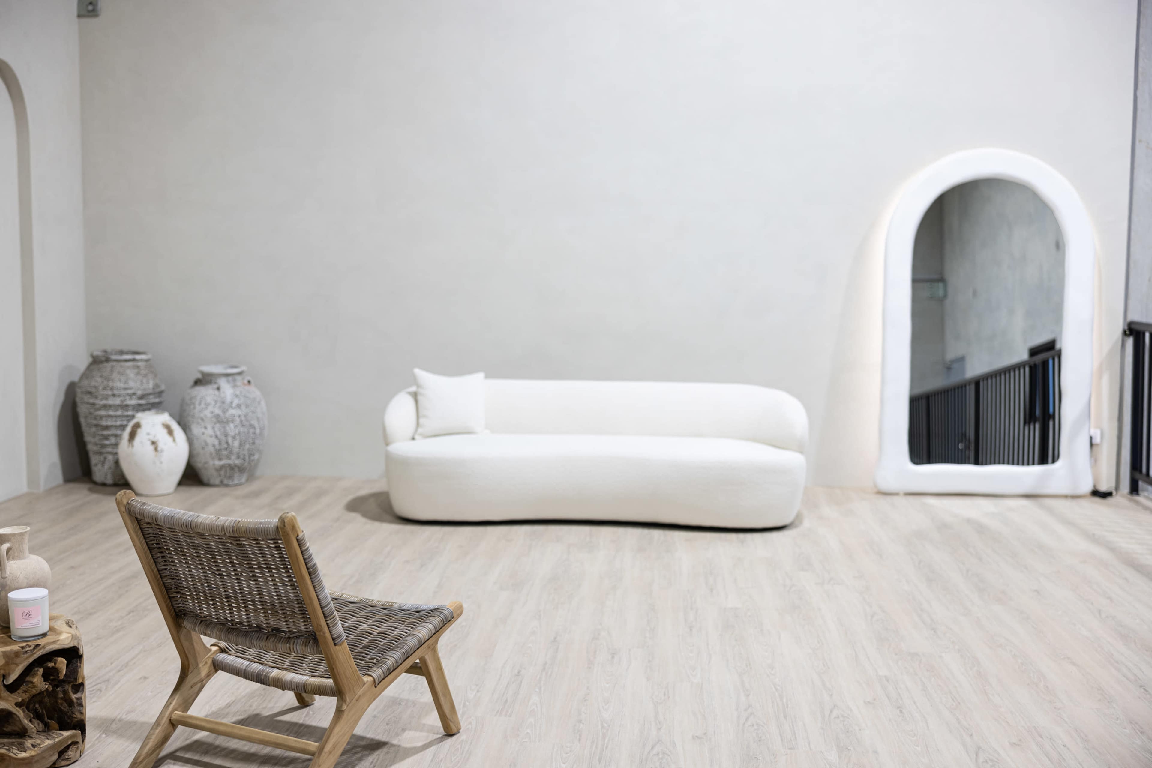 The image shows a minimalist interior space with a white sofa, a wooden chair, decorative vases, and an arched mirror against a light-colored wall.