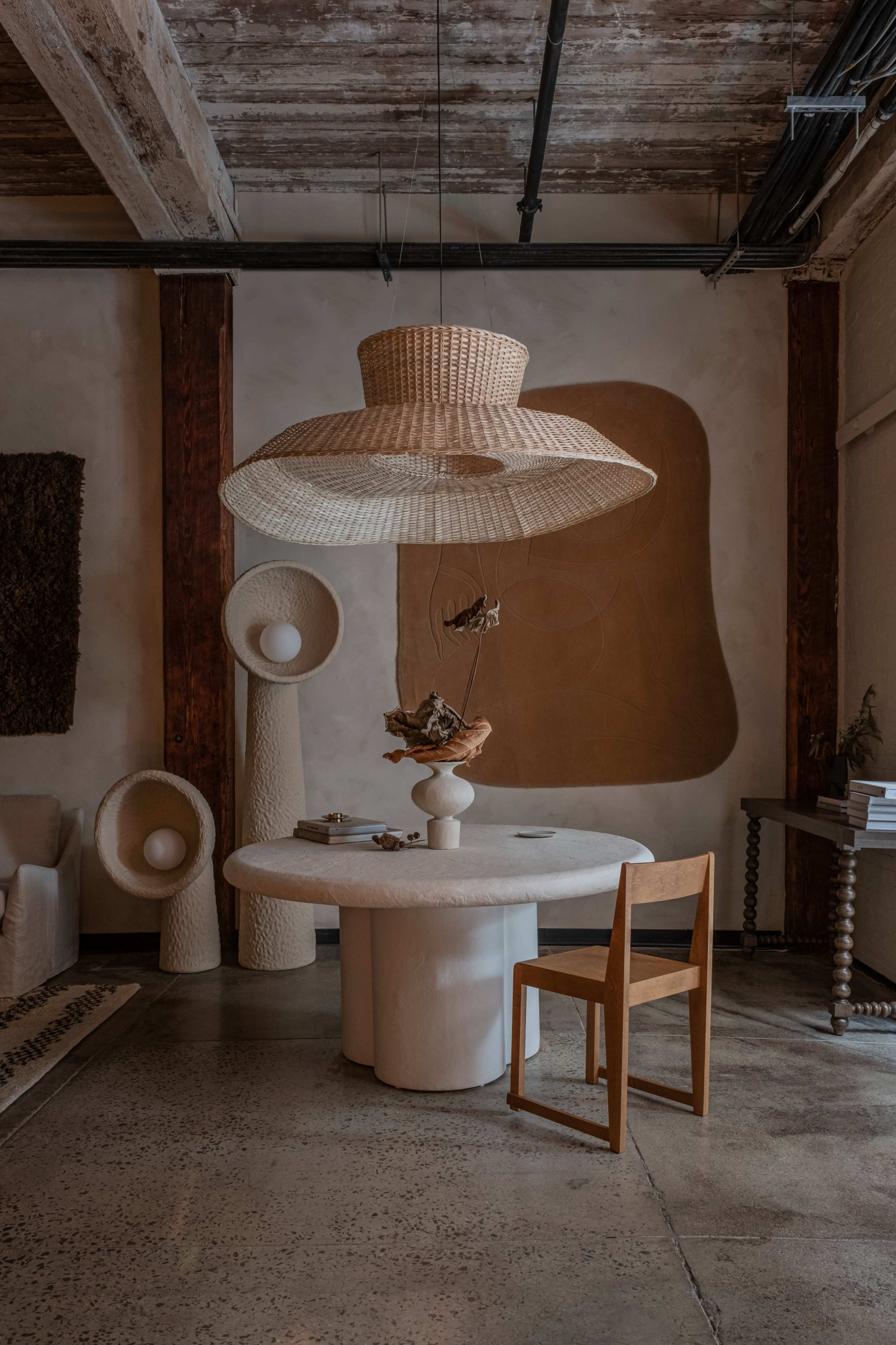A modern dining area with a round, white table, a wooden chair, and a large woven pendant light overhead, surrounded by textured wall art and decorative sculptures.