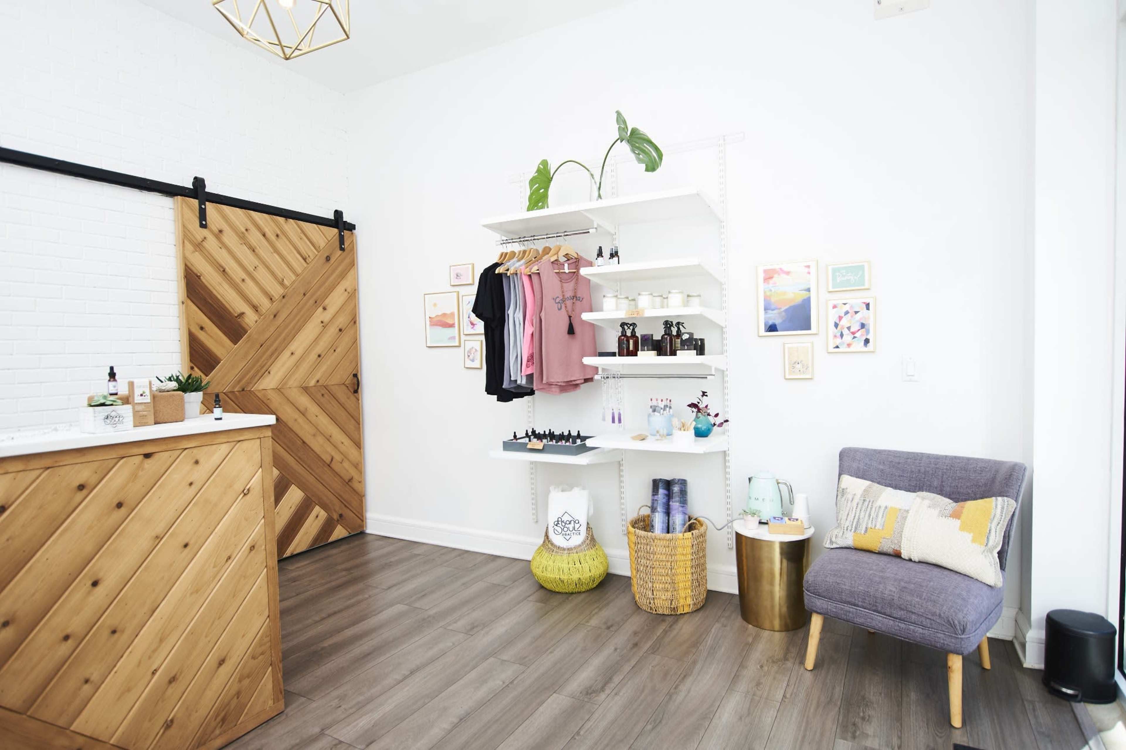 The image shows a retail space featuring a wooden reception counter, shelves displaying products, and a comfortable chair next to decorative items against a white brick wall.