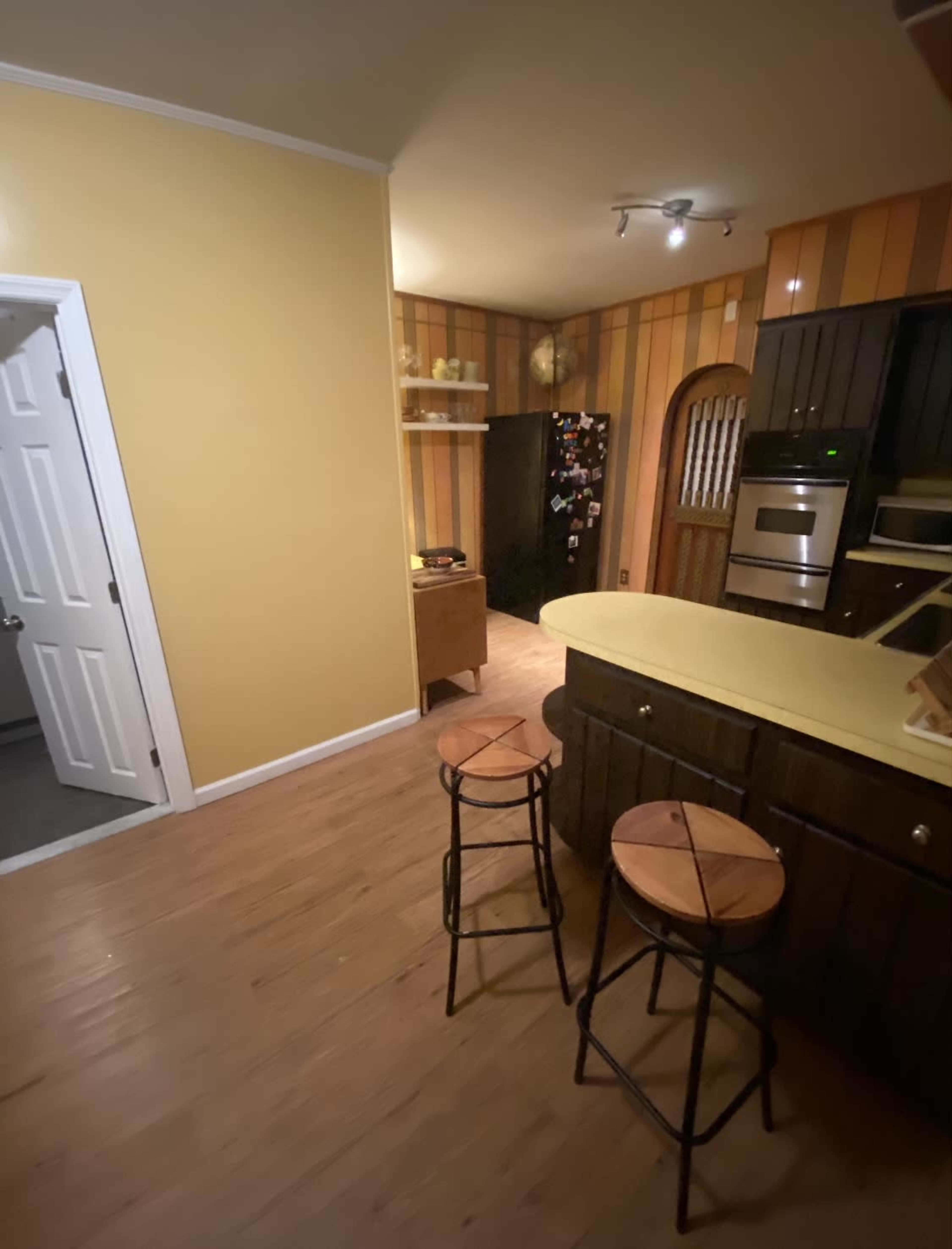 A kitchen features yellow walls, wooden stools at a counter, and appliances along with a partially visible pantry.