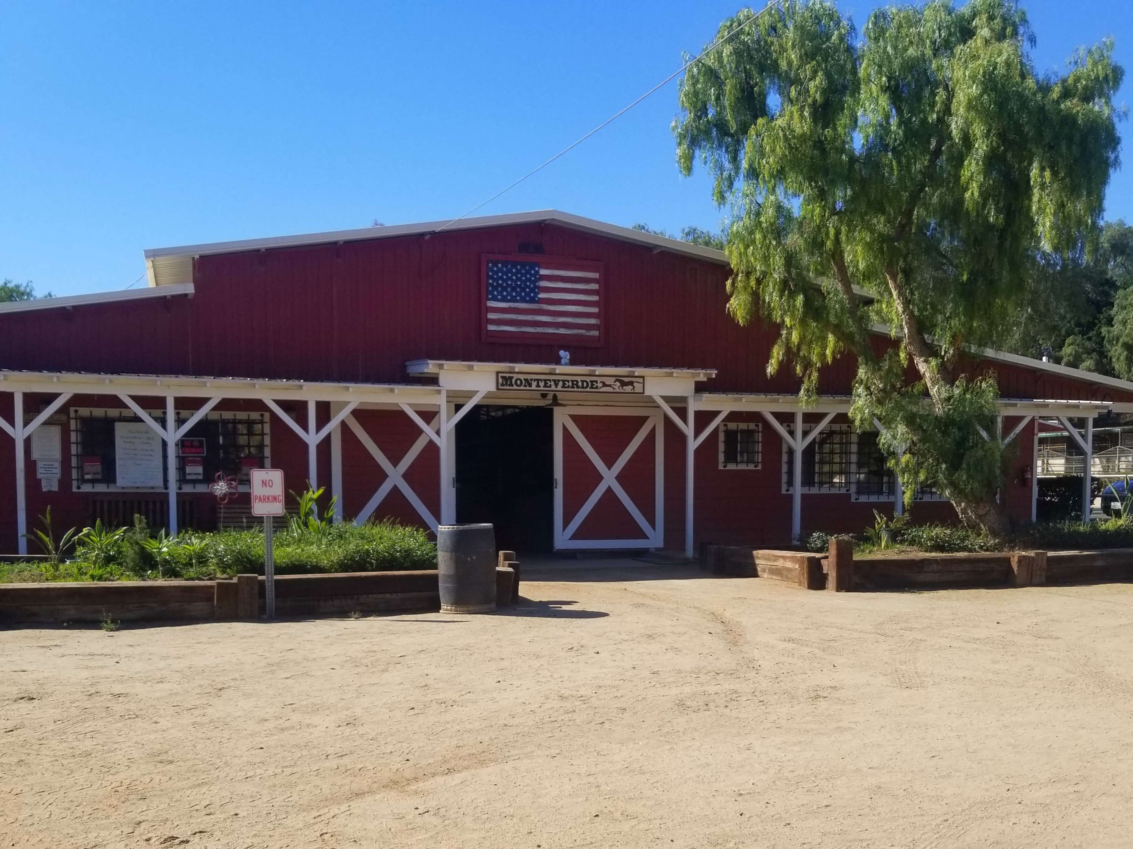 A red barn with a flag on the front is surrounded by greenery and features a gravel path leading to the entrance.