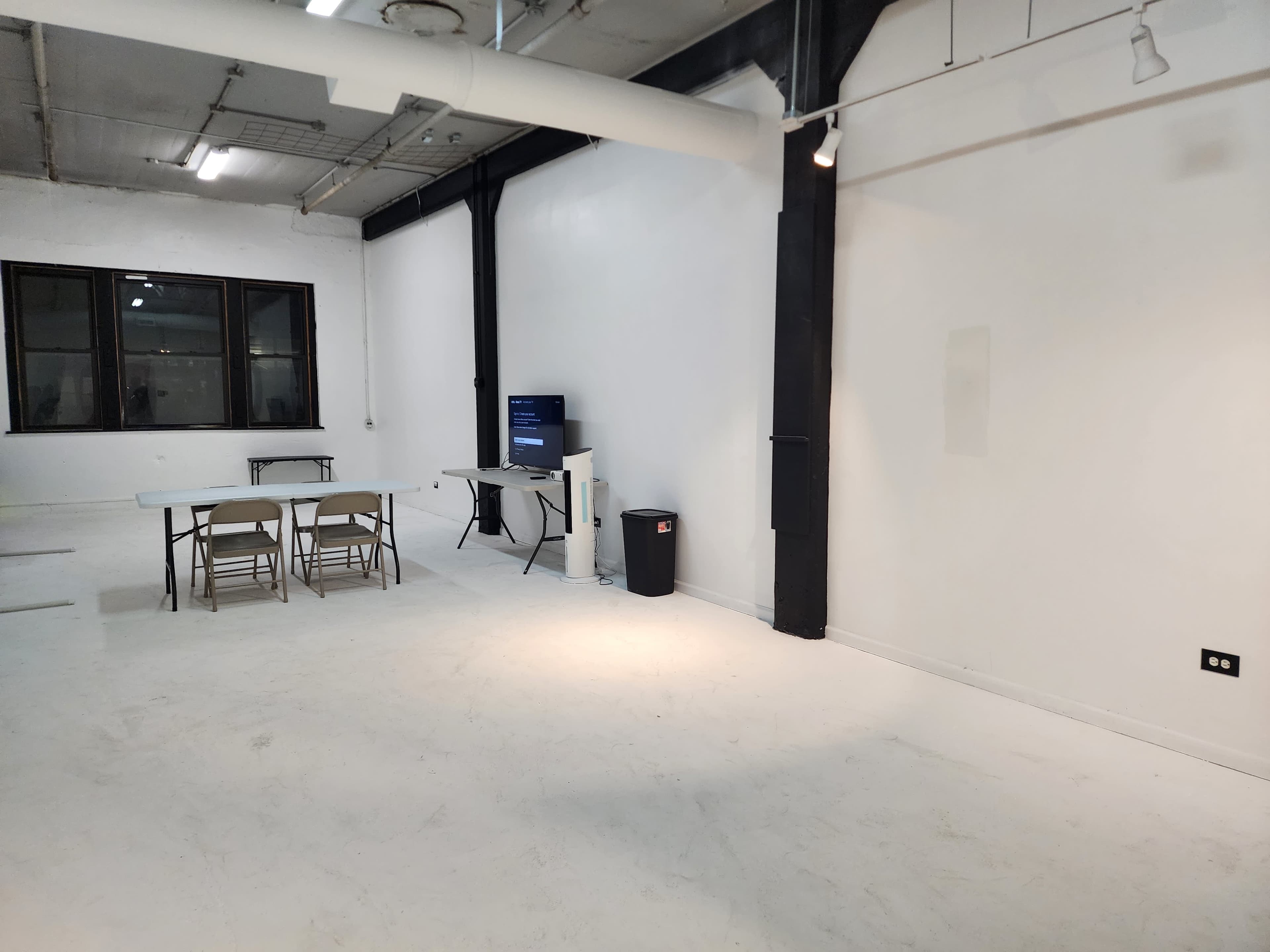 The image shows an empty room with white walls, a table and chairs, a television on a stand, and a trash can.