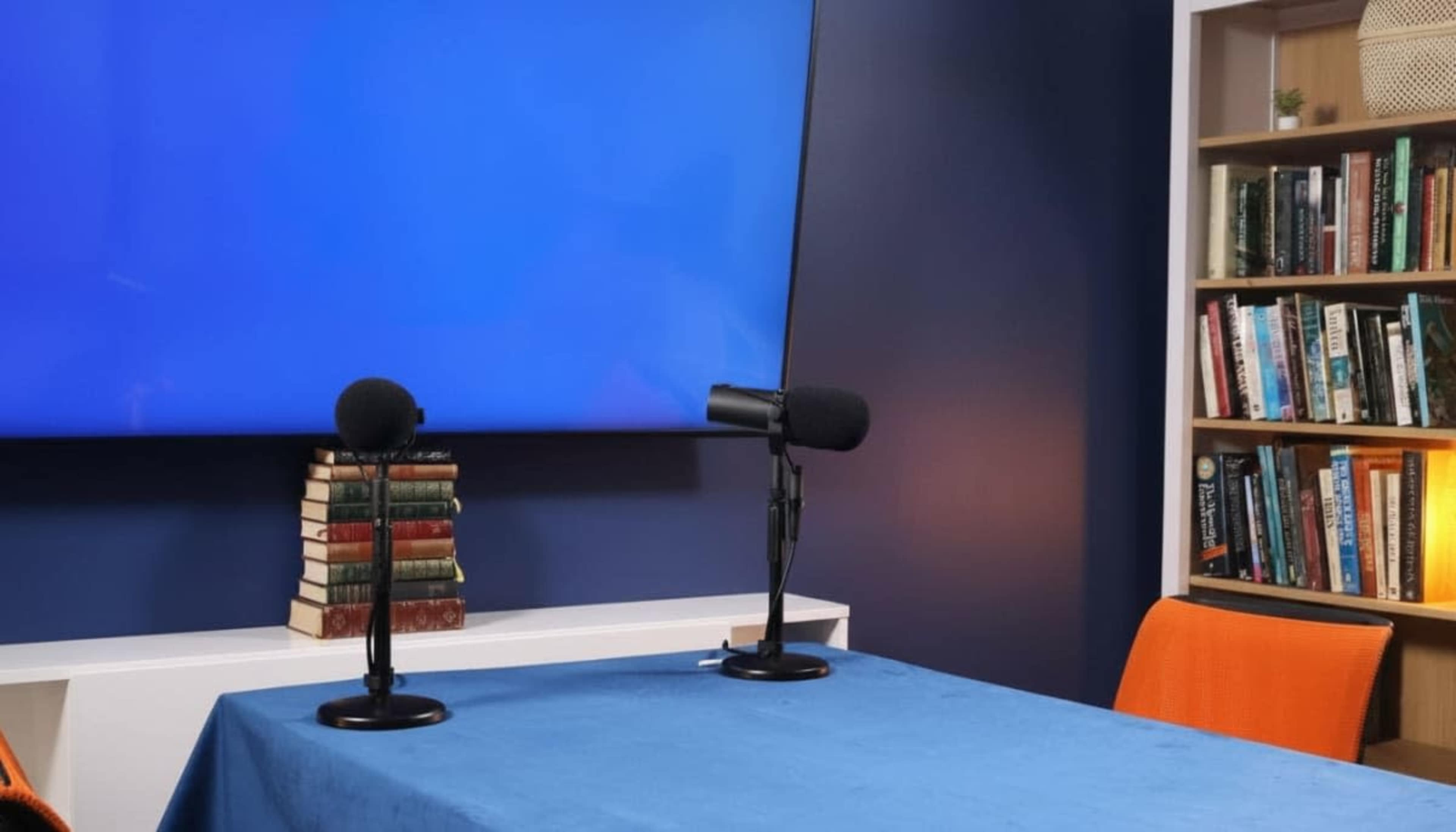 The scene features a table set with two microphones in front of a large blue screen, accompanied by a bookshelf filled with books and two orange chairs.