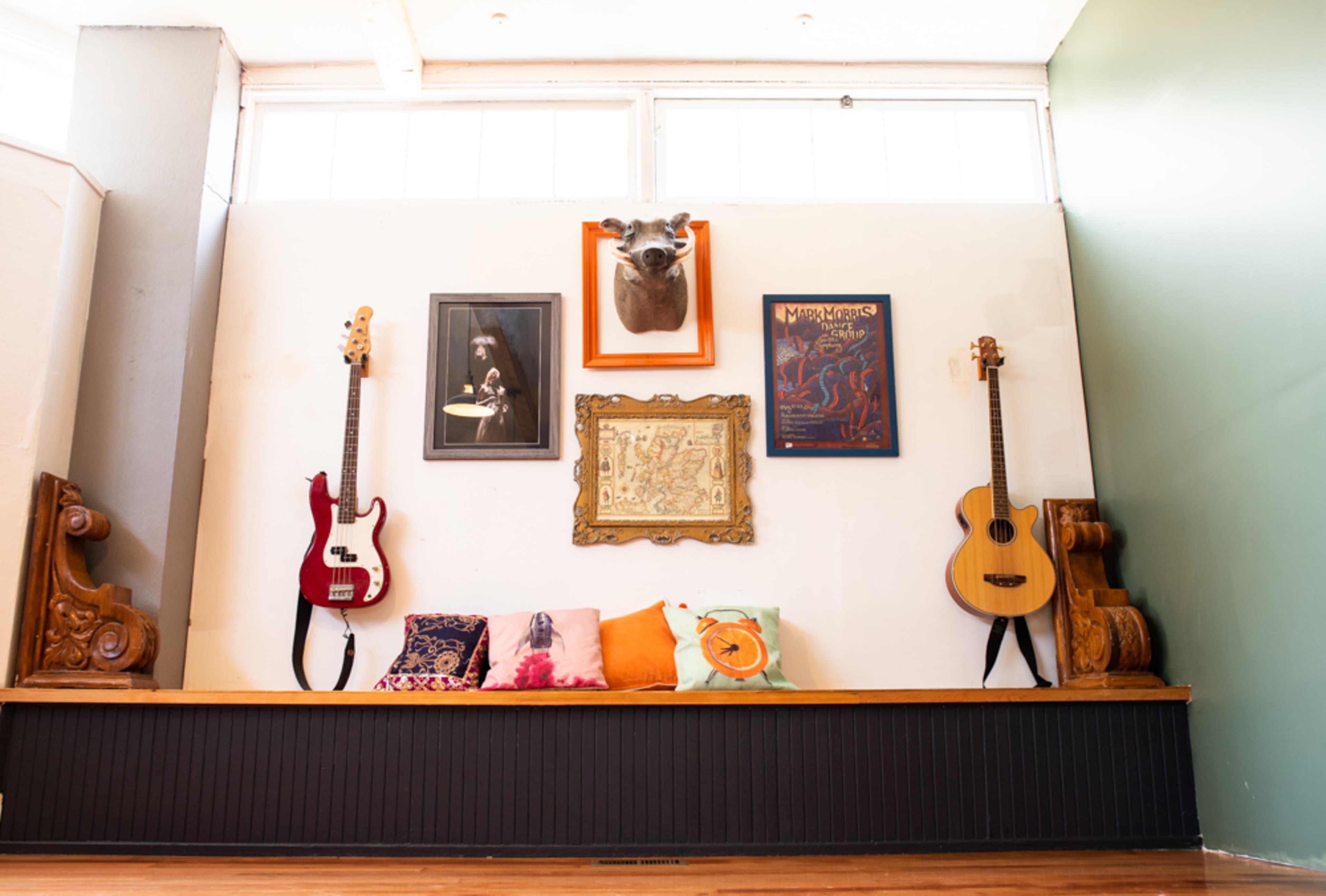 The scene features a decorative wall with two guitars, framed artwork, a mounted animal head, and a collection of colorful cushions arranged on a wooden bench.