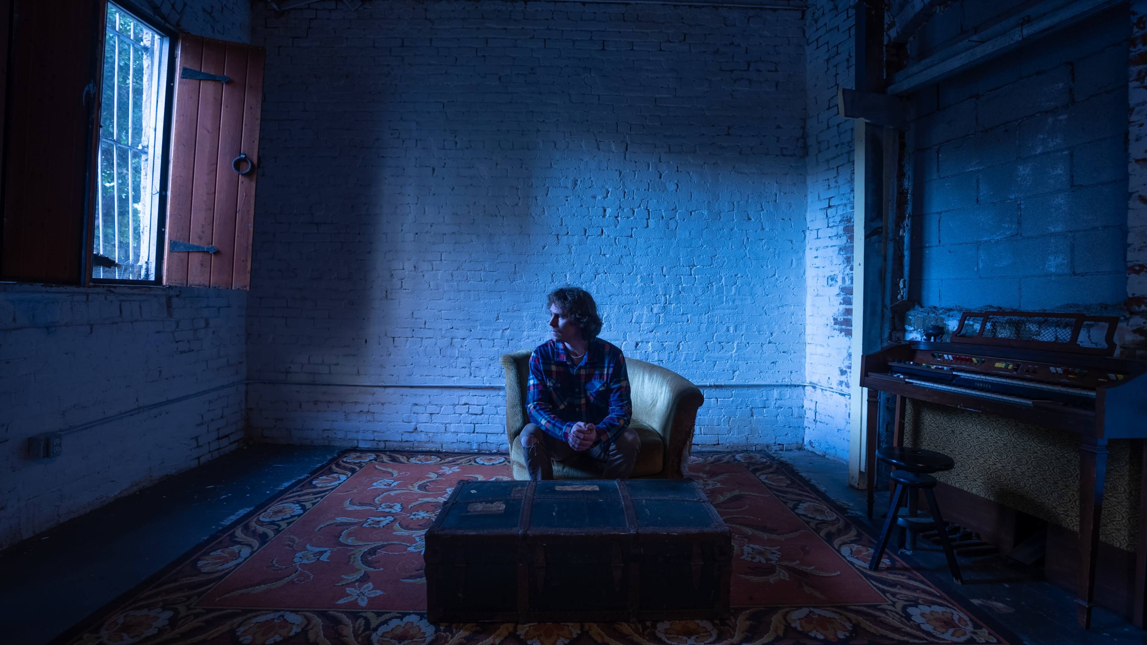 A person sits on a yellow chair in a dimly lit room with exposed brick walls and a vintage rug, beside an old piano and a wooden trunk.