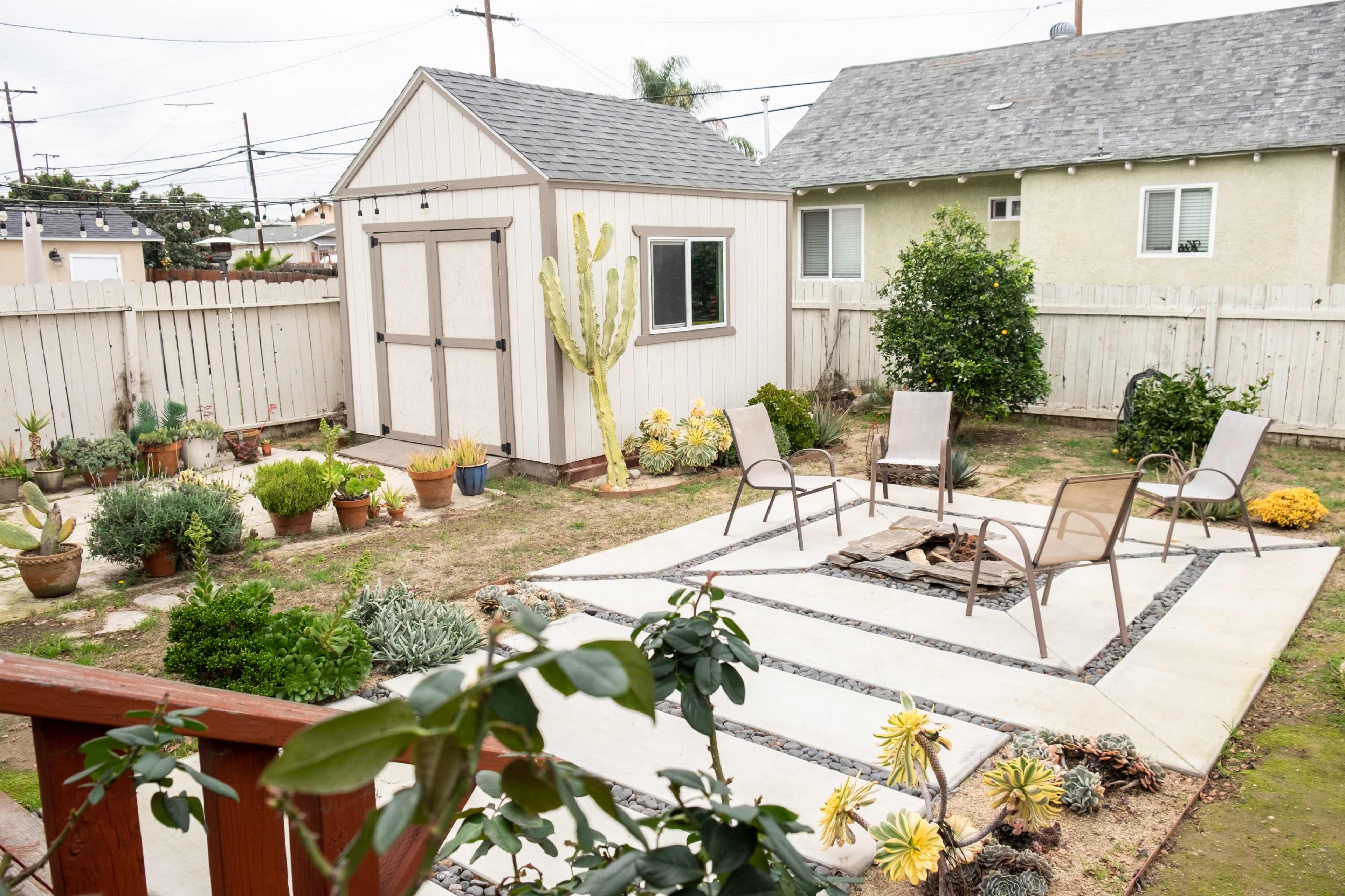 A backyard with a small shed, a fire pit area surrounded by chairs, and various plants and cacti arranged around a concrete pathway.