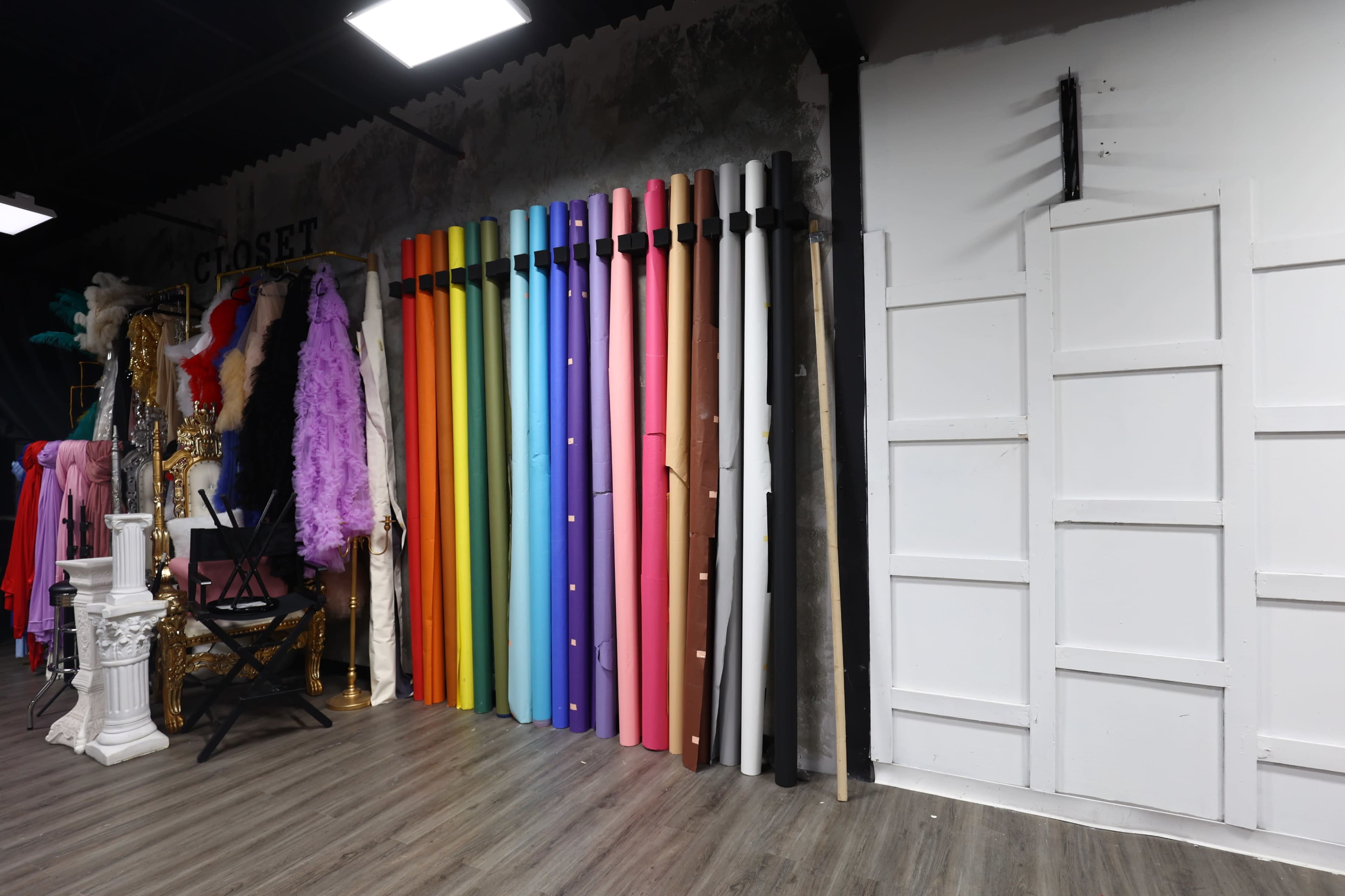 A wall displays a variety of rolled backdrop papers in multiple colors alongside a collection of garments and props in a studio setting.