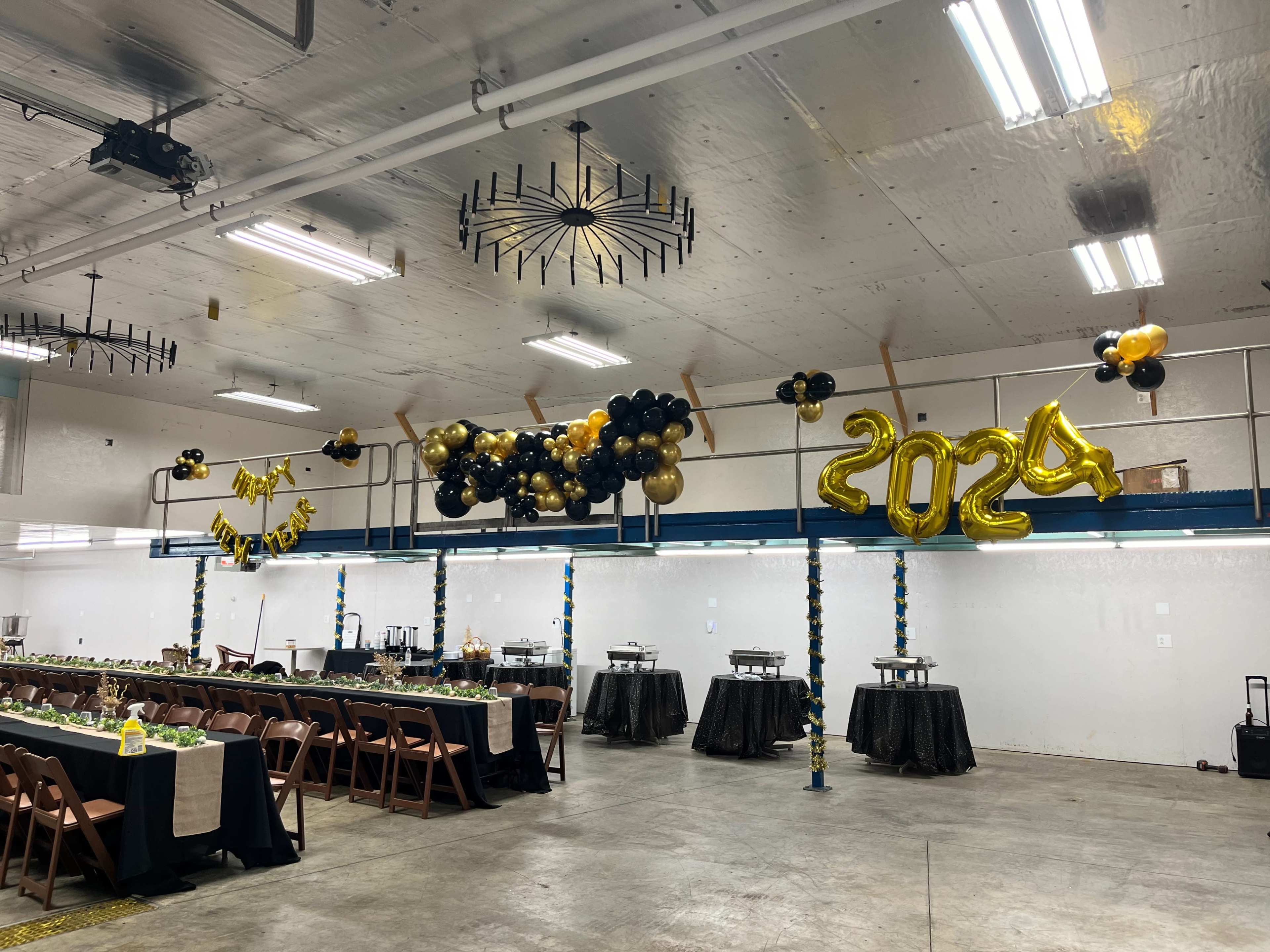 The image shows a decorated indoor space with two long banquet tables set for a celebration, adorned with black and gold balloons and a "2024" balloon display.
