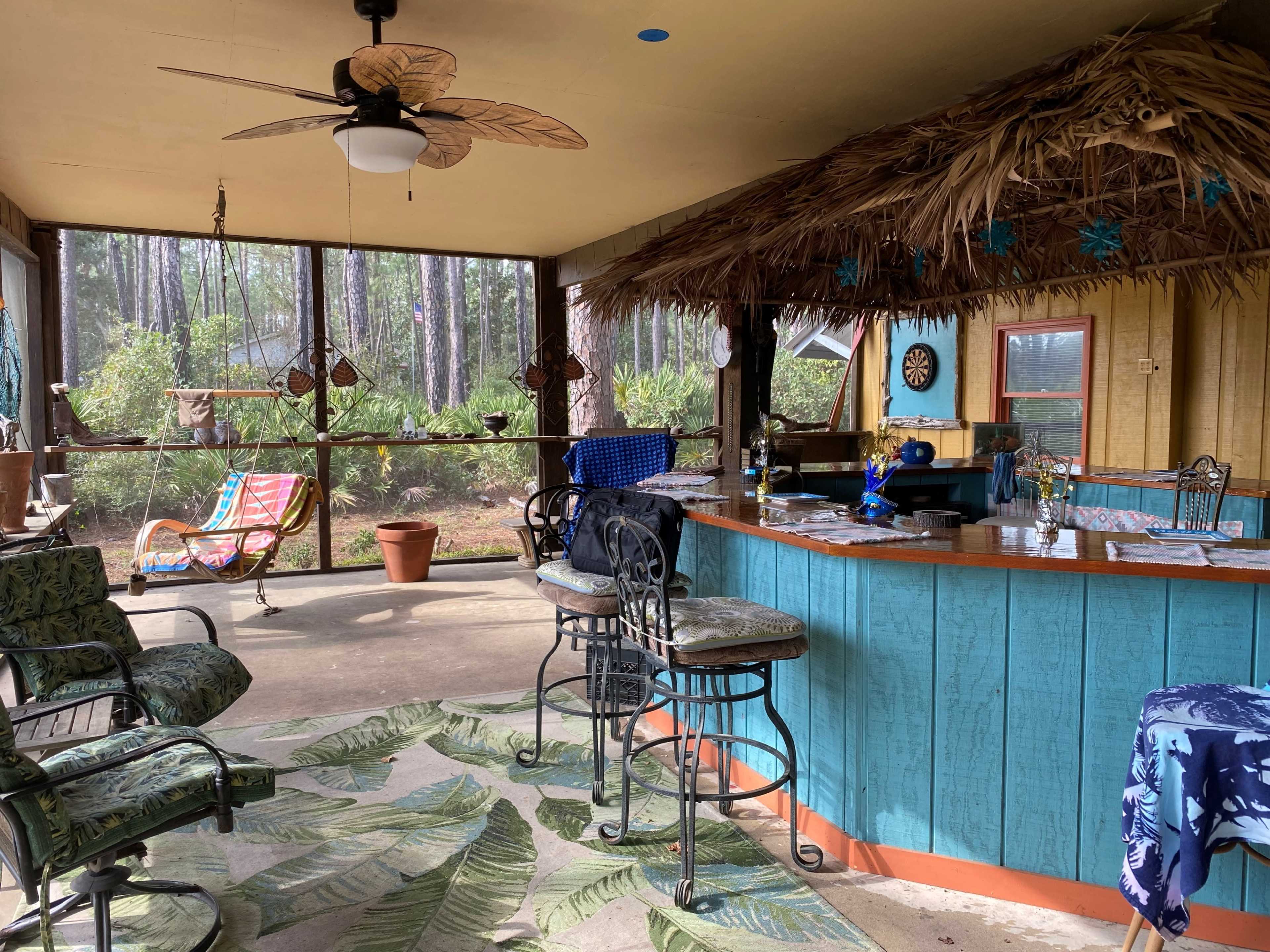 A screened-in porch featuring a bar with high stools, tropical-themed decor, and seating arrangements under a thatched roof.