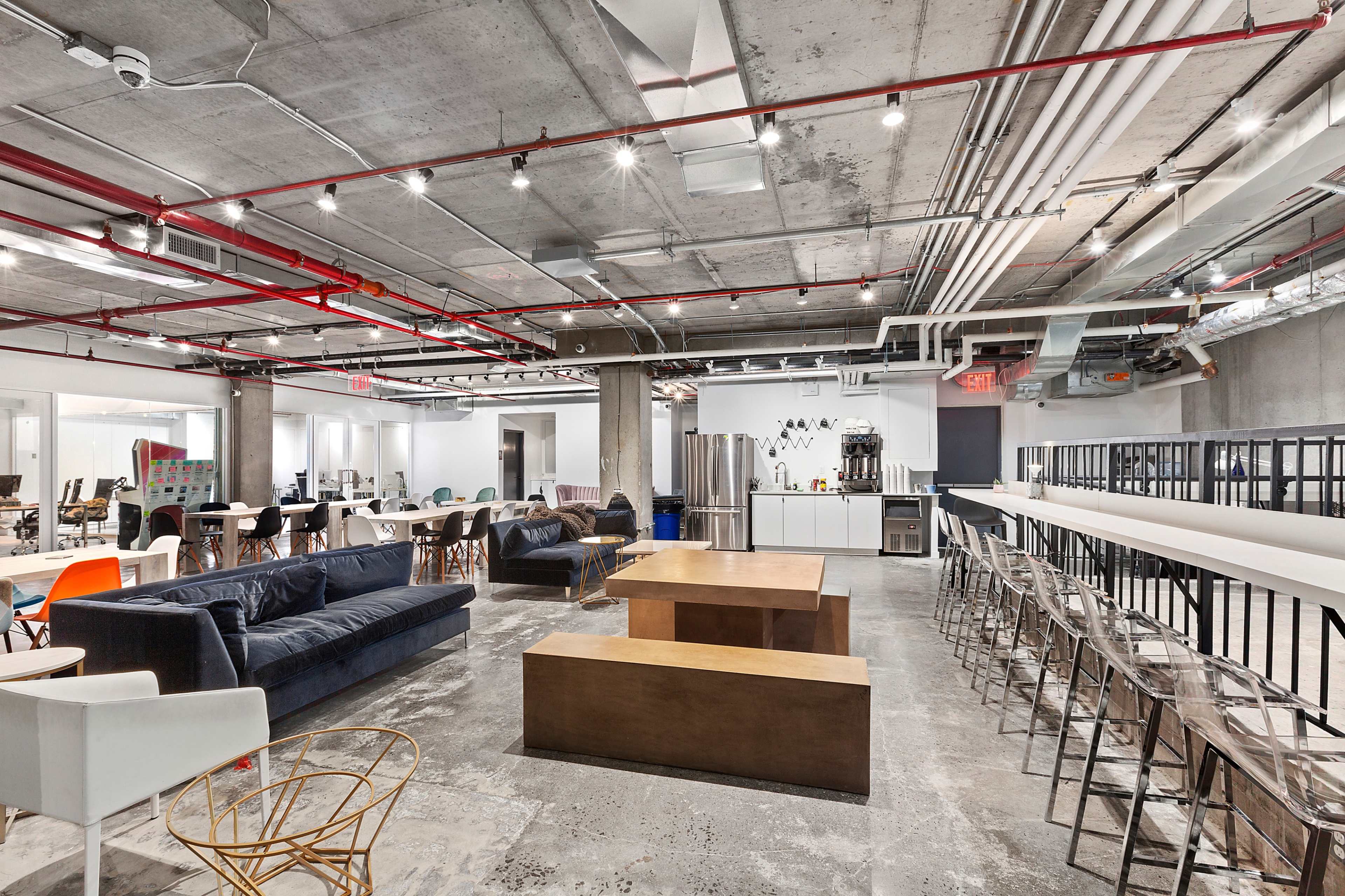 A modern office space featuring various seating areas, including a lounge with sofas, tables, and a kitchenette area with appliances.