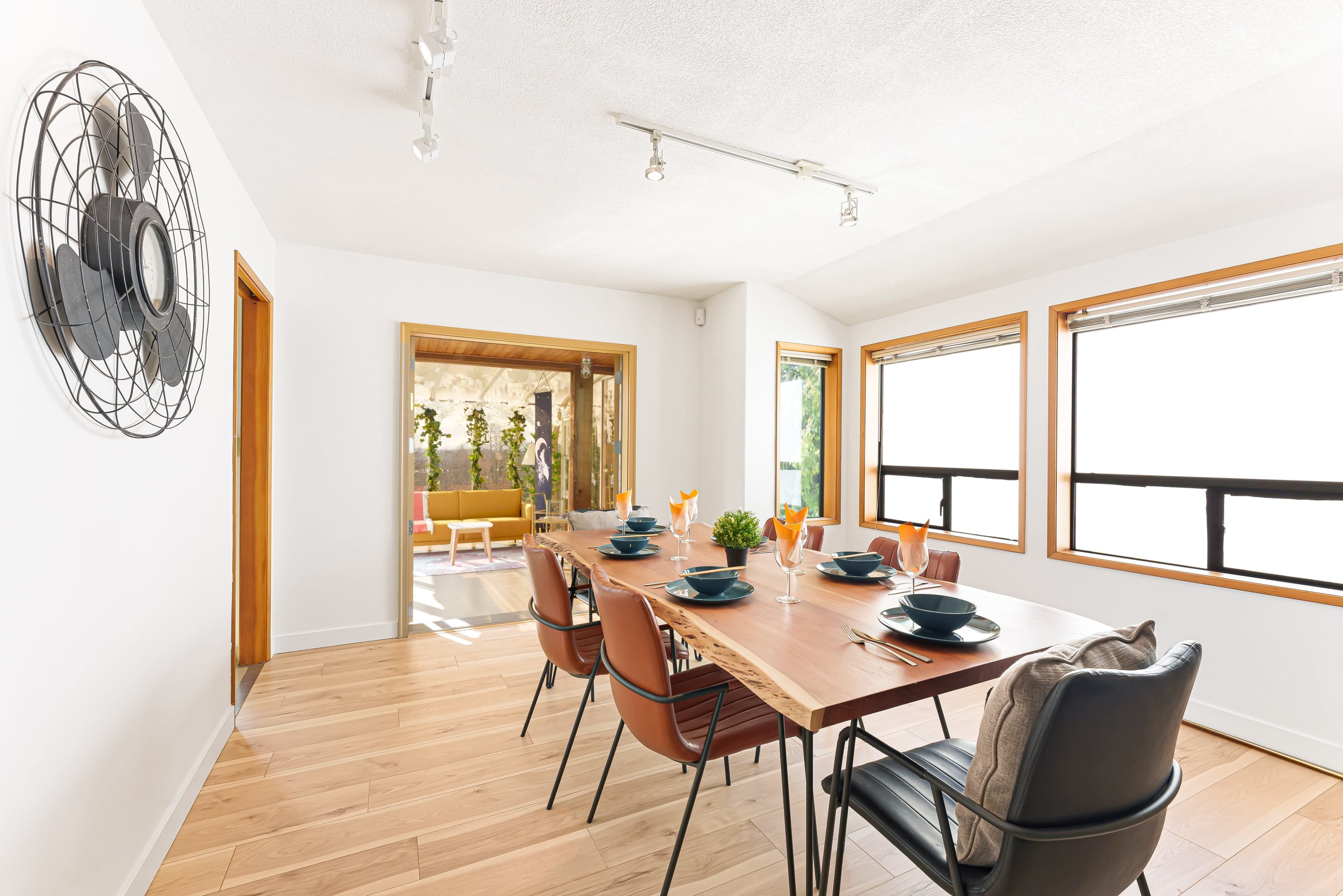 A bright dining area features a wooden table set for six with modern chairs and large windows that provide natural light.